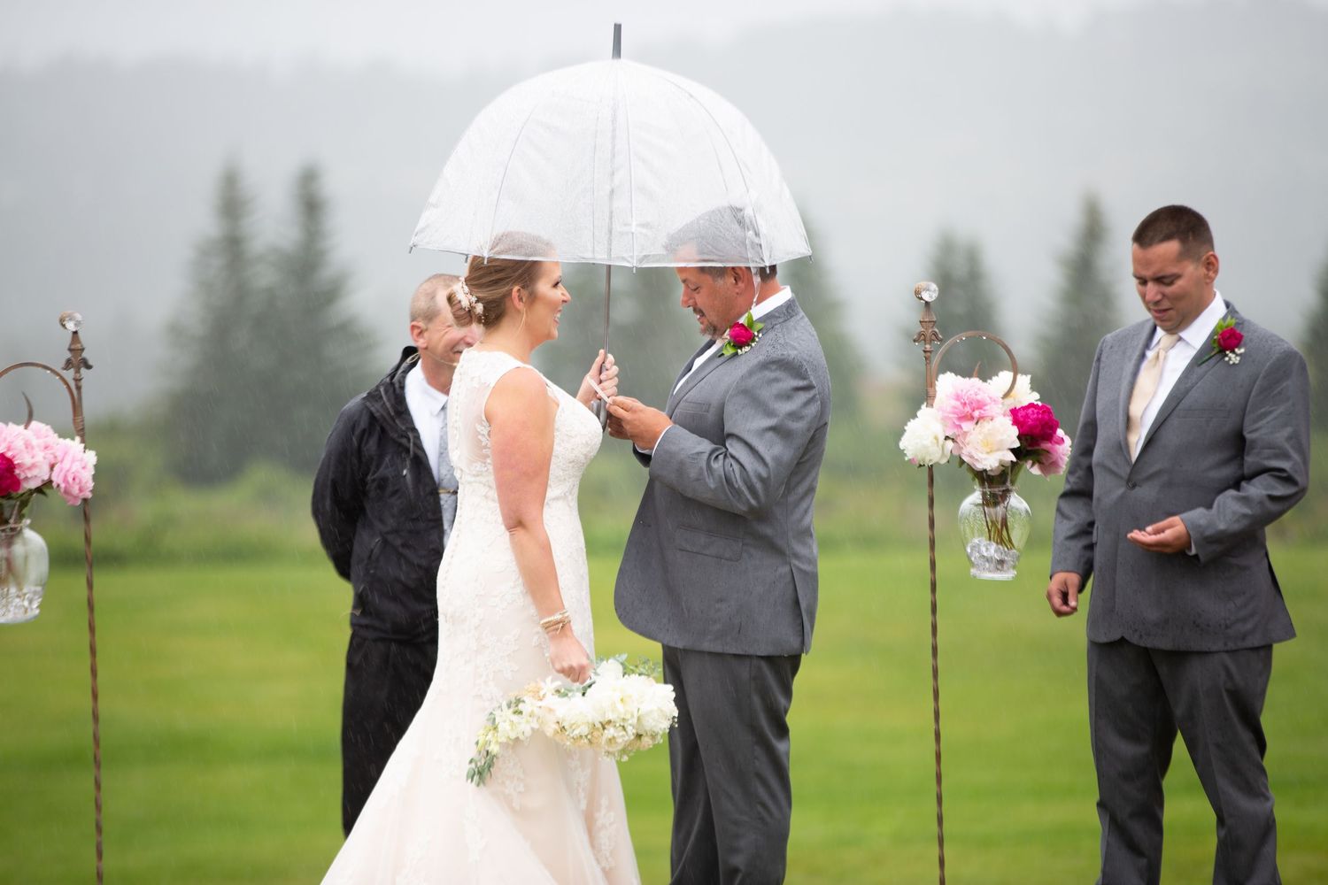 Wedding ceremony on a rainy day with attendees holding white umbrellas on a green lawn.