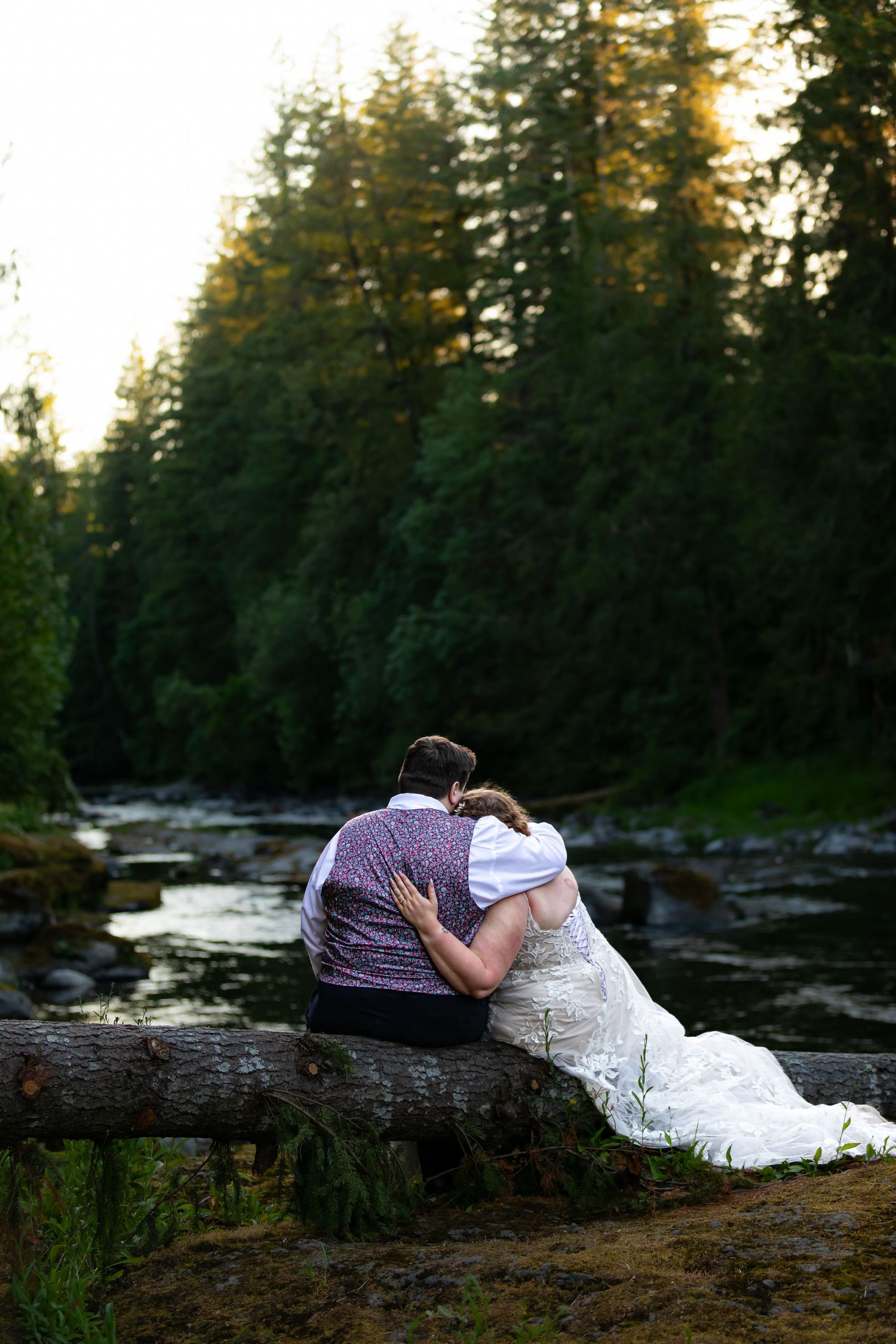 Wedding couple relaxes on fallen log by river during golden hour photography session.