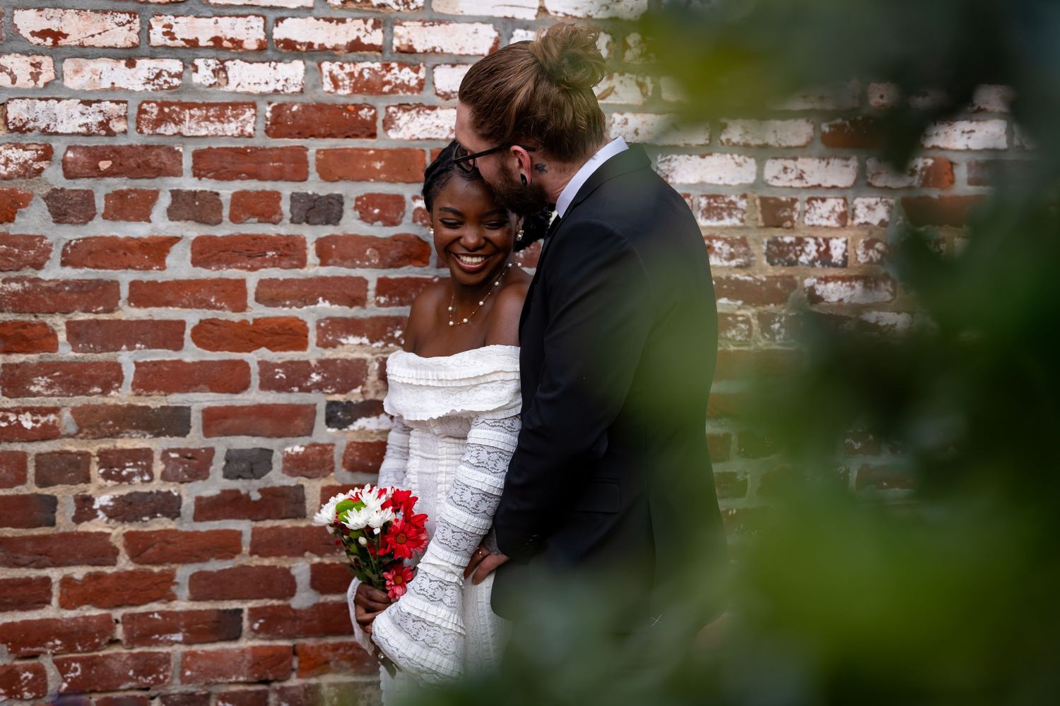 A tender embrace photographed through foliage against a brick wall background.