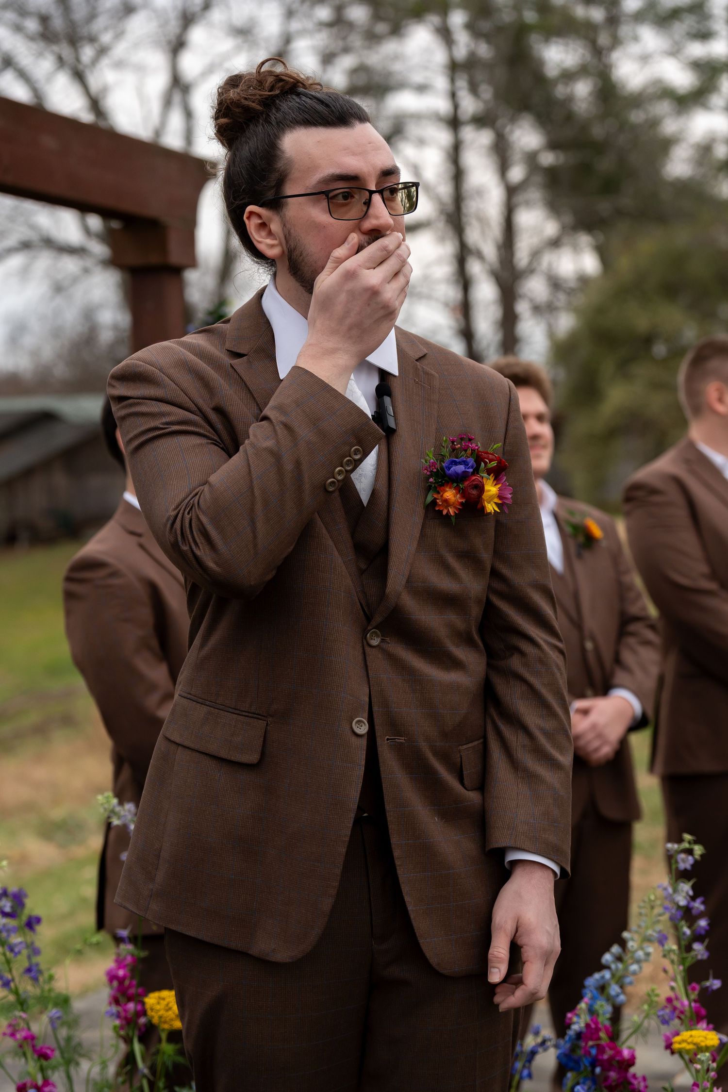 Groomsmen in brown suits react emotionally during outdoor wedding ceremony with colorful flowers.