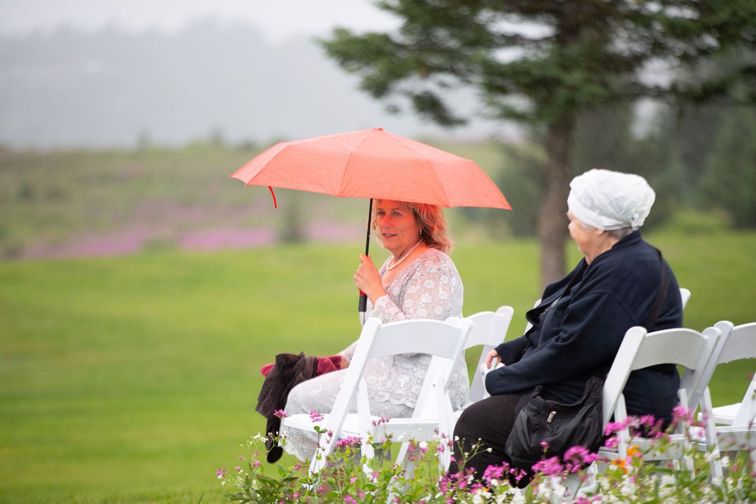 Two guests sit in white chairs on a lawn during a rainy outdoor ceremony with one holding a coral umbrella.