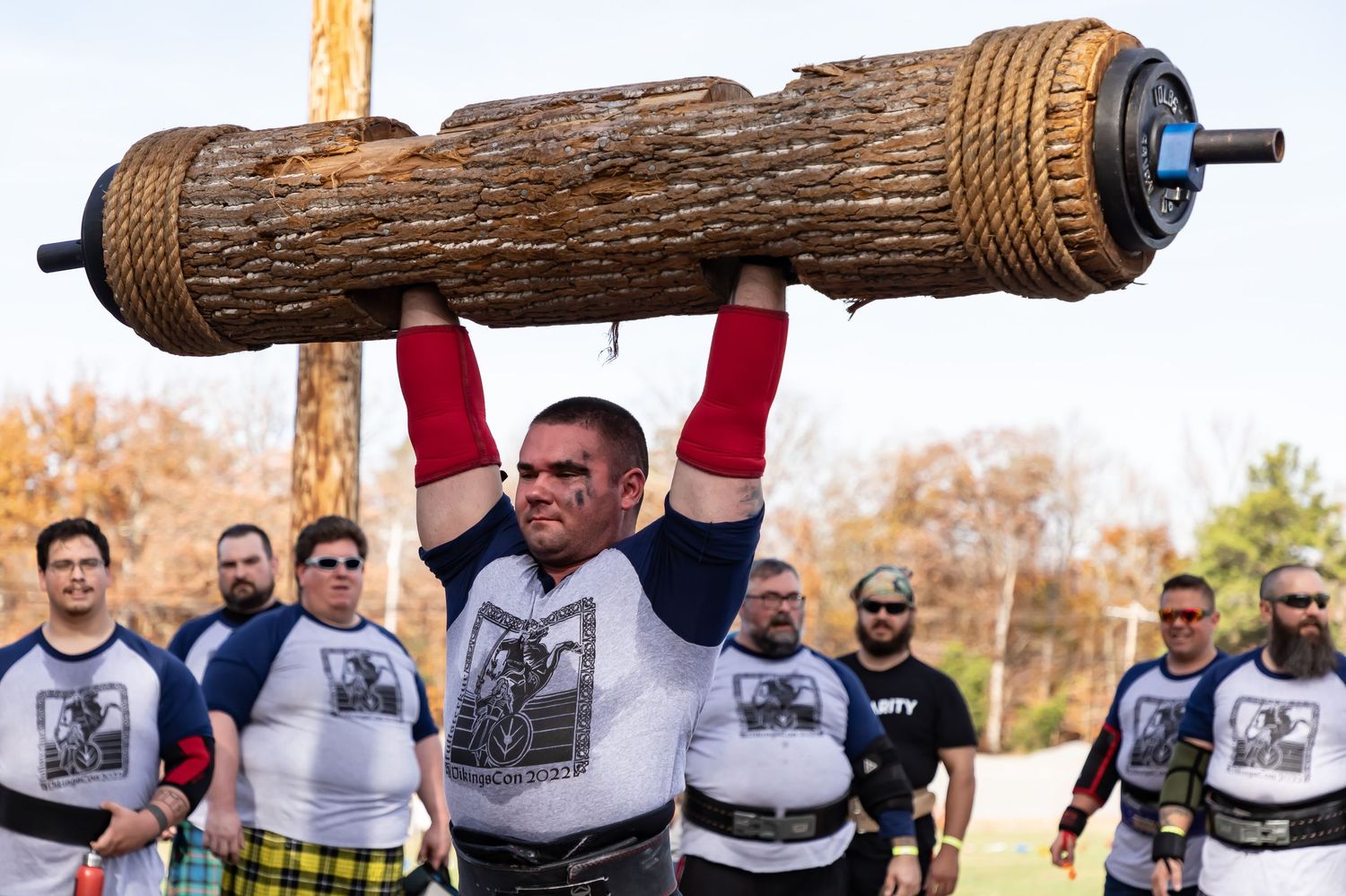 Strongman competitors in matching uniforms prepare to lift decorated wooden log.