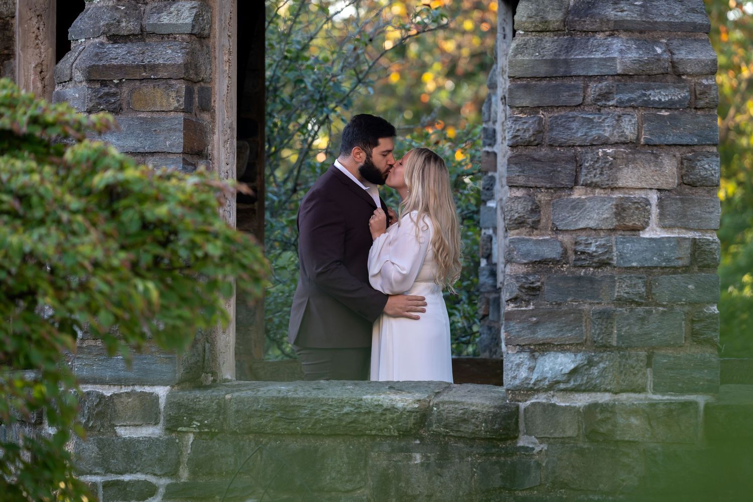 Romantic couple poses between stone pillars in garden during wedding photoshoot.