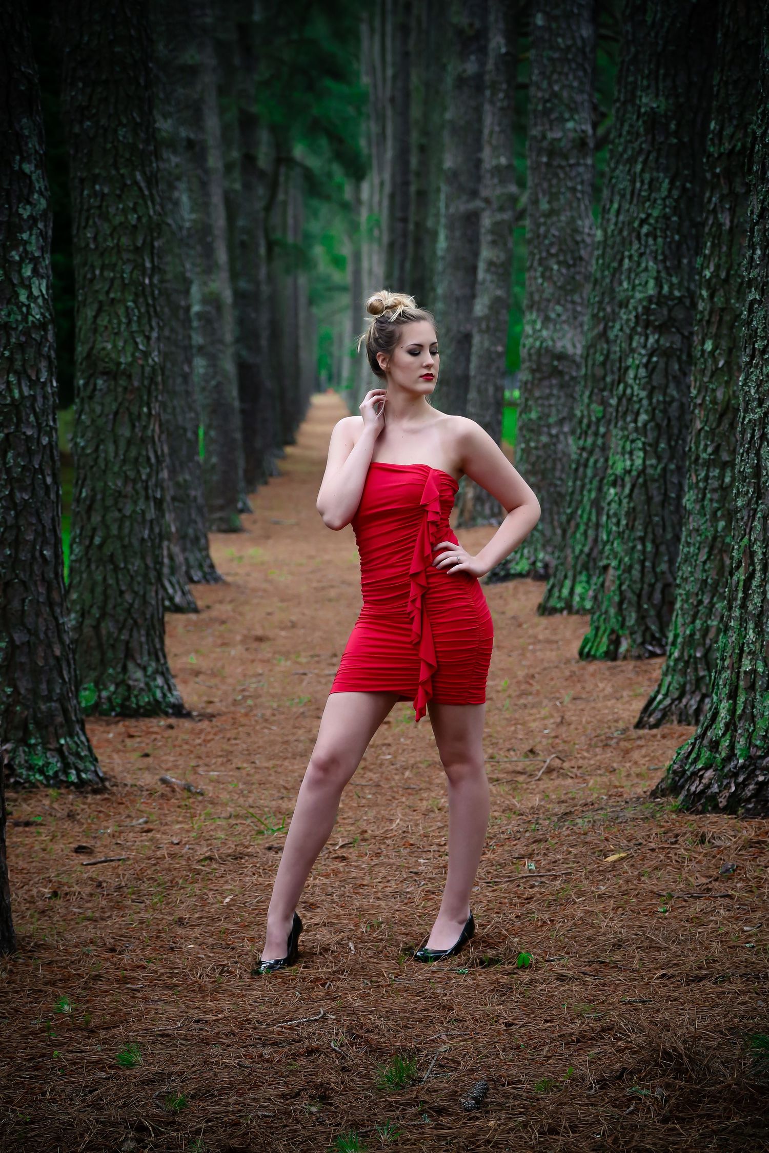 Dynamic poses in a red dress among rows of tall pine trees.
