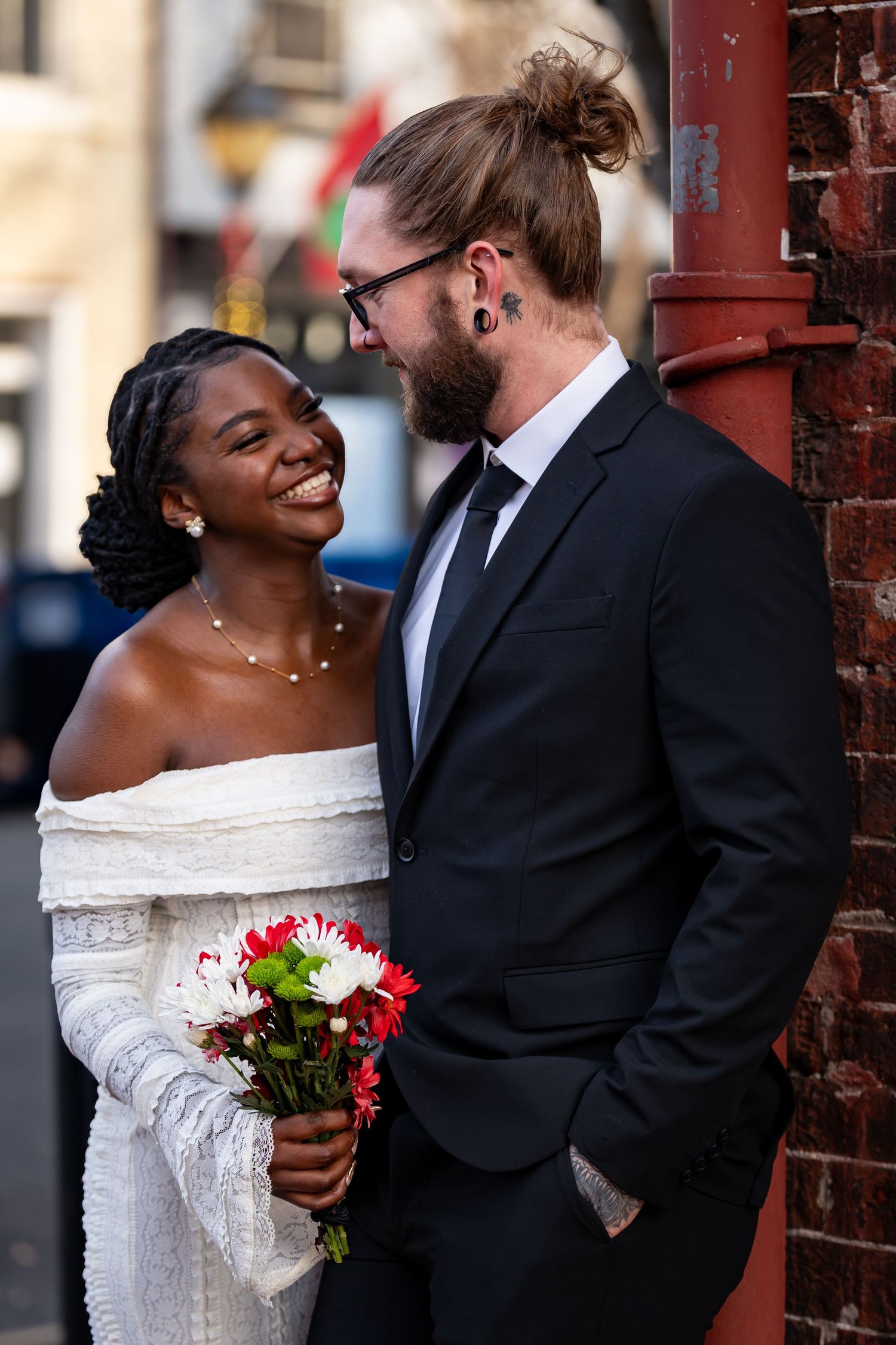 A joyful couple shares an intimate moment against a brick wall while holding a wedding bouquet.