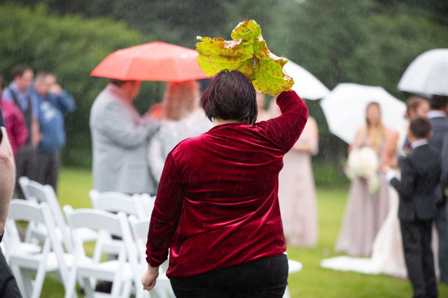 Wedding guests in formal attire stand with umbrellas during an outdoor rainy ceremony.