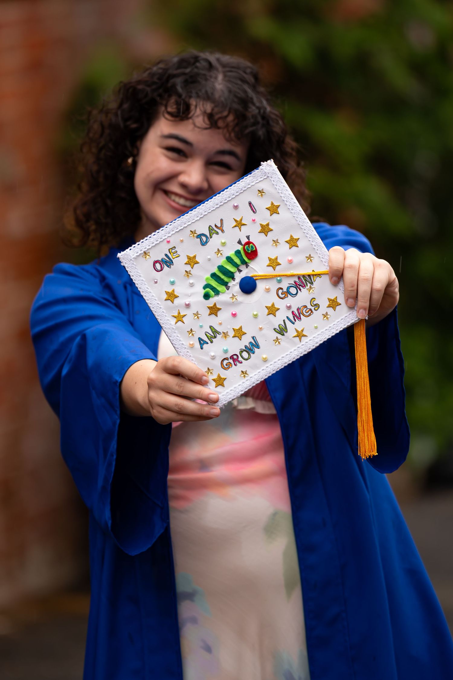 Graduate displays decorated graduation cap with stars and text while wearing blue commencement gown.