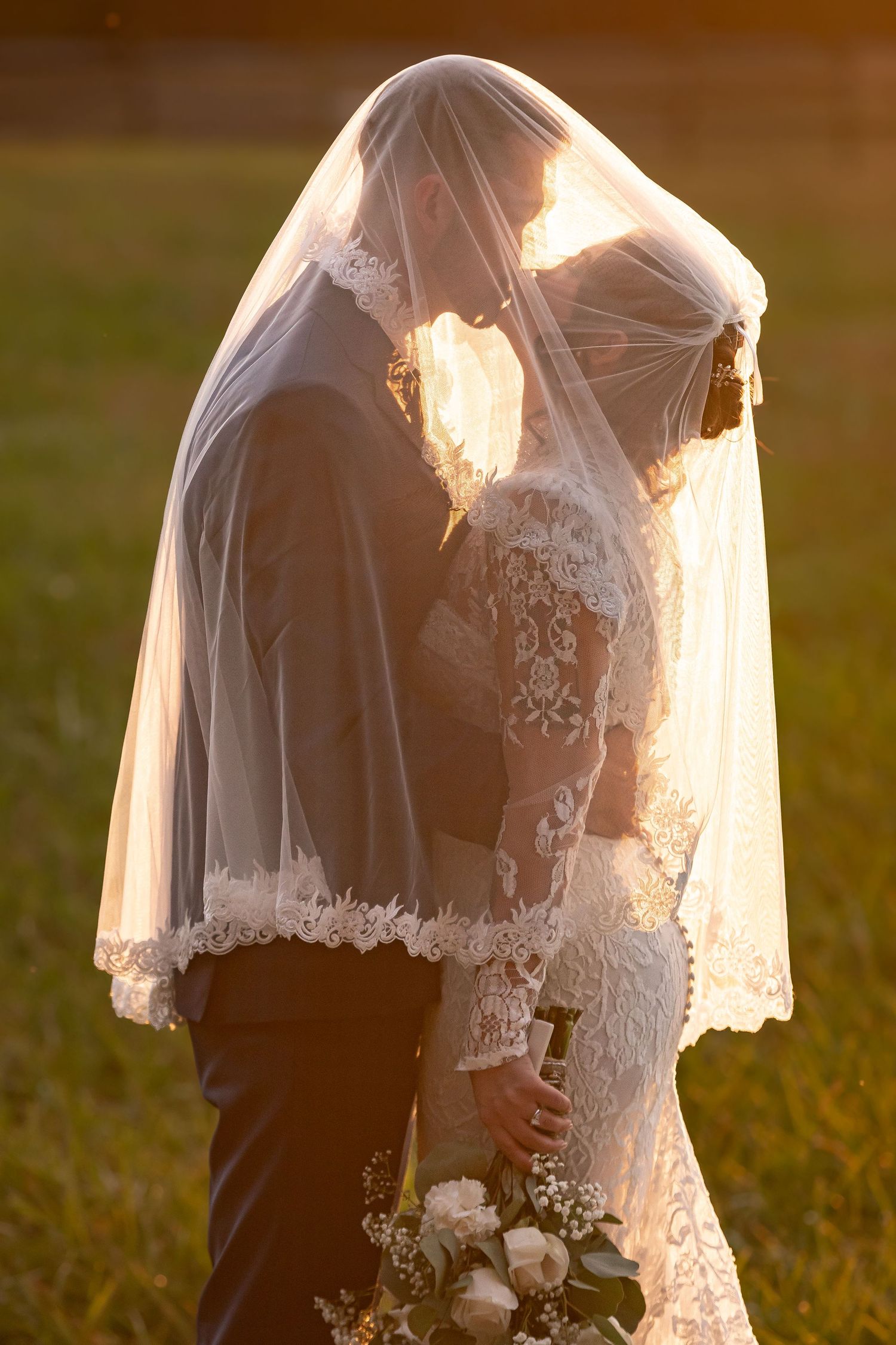 Romantic sunset portrait of couple embracing under sheer wedding veil.