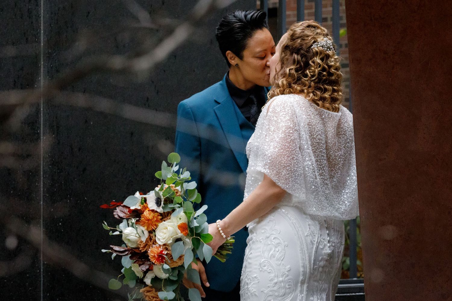 Two brides, one in white lace and one in a blue suit, with a colorful floral bouquet during a First Look at an intimate wedding ceremony.