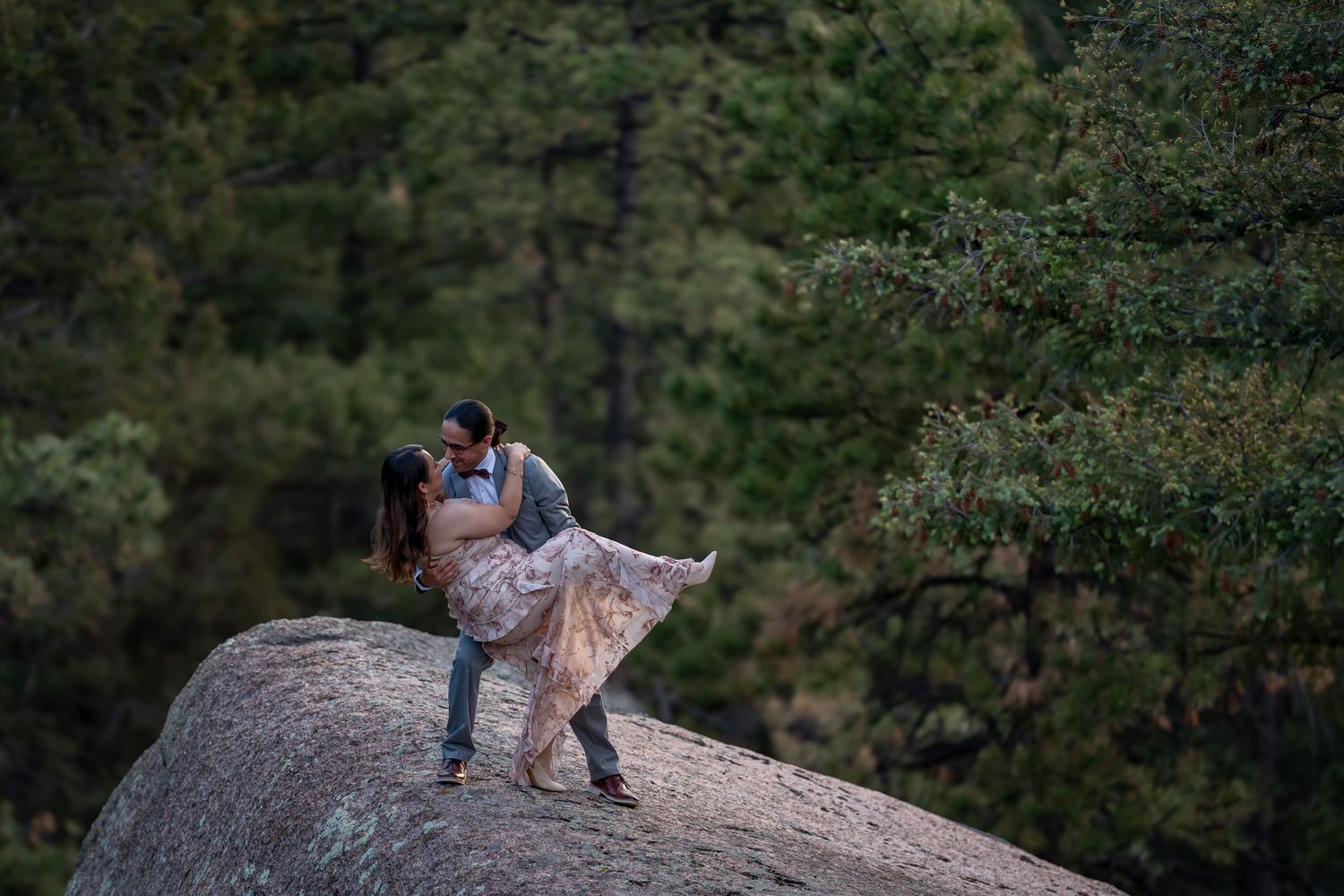 Romantic couple with woman in a floral dress posing on large boulder in wooded mountain setting in the Rocky Mountains in Sedalia, Colorado.