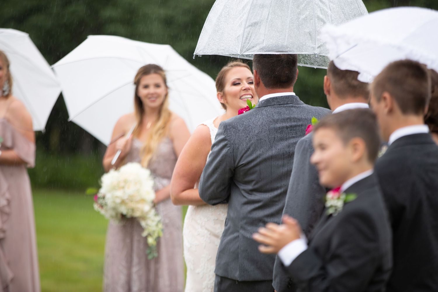 Wedding party holding white umbrellas outdoors in a garden setting during light rain.