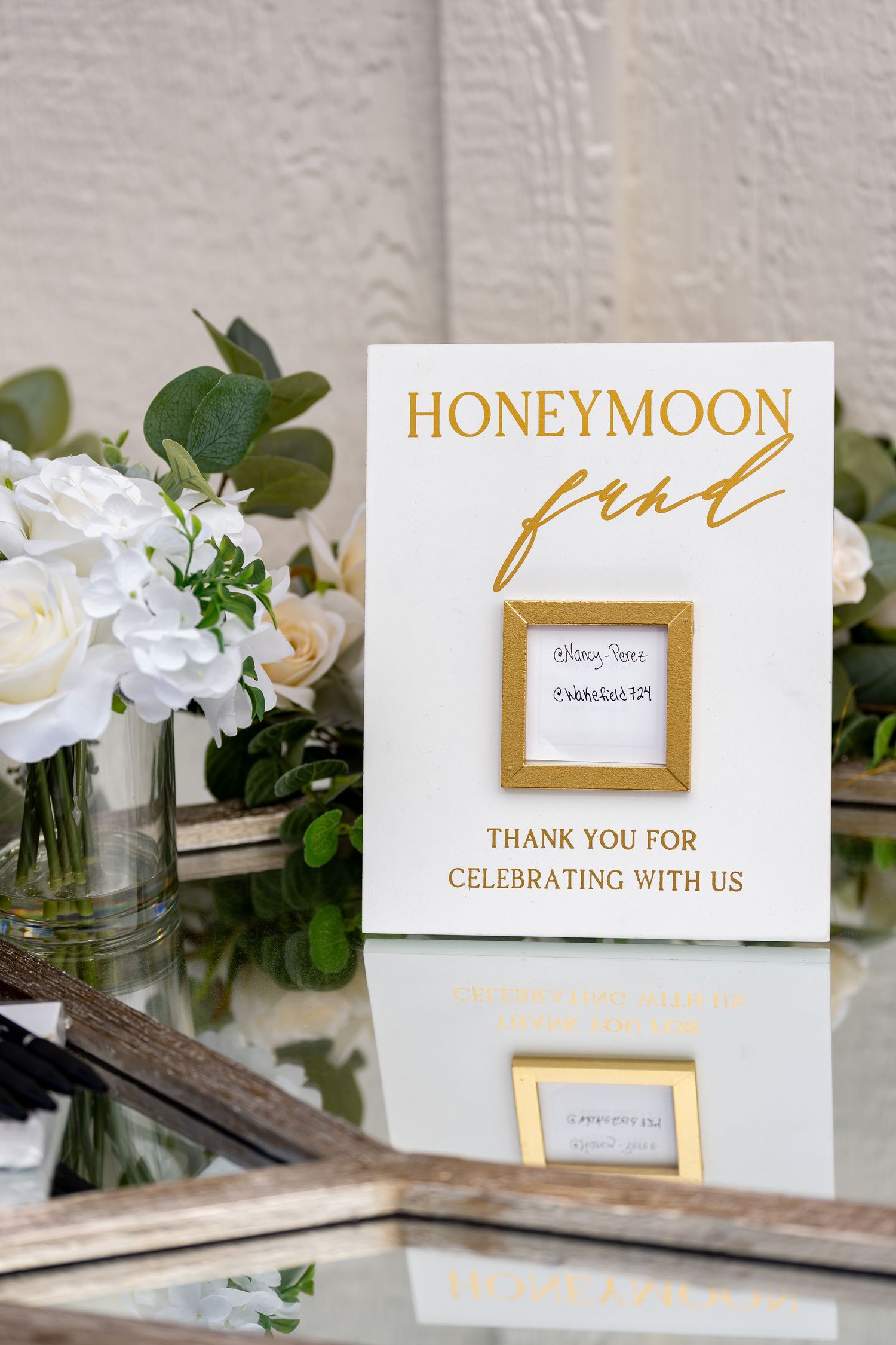 Gold and white honeymoon fund sign with floral arrangements and greenery.