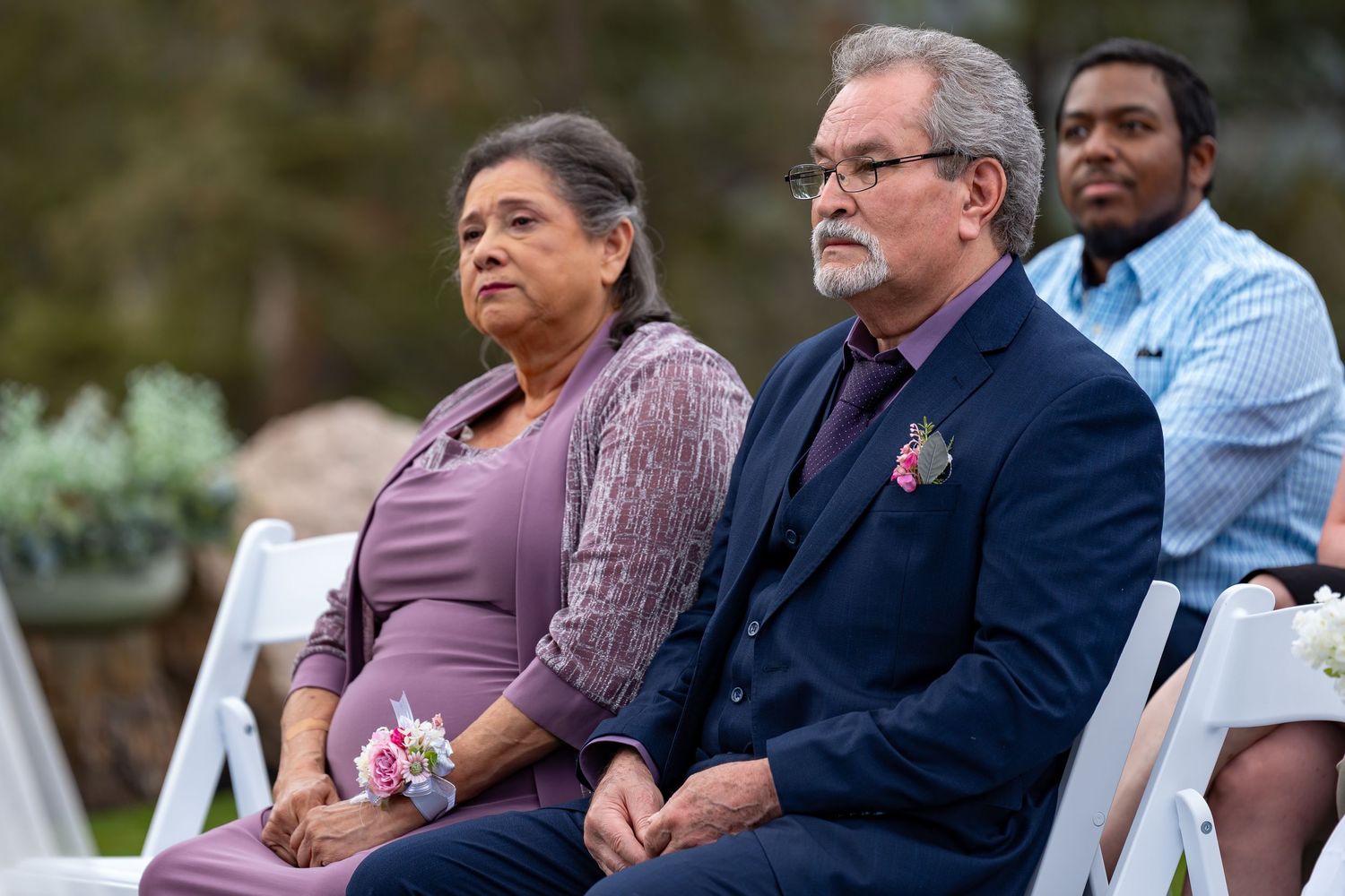 Attendees in formal attire observe an outdoor wedding ceremony from white chairs.