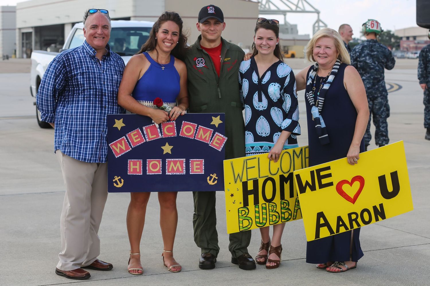 Family and friends hold colorful welcome home signs at military base reunion.