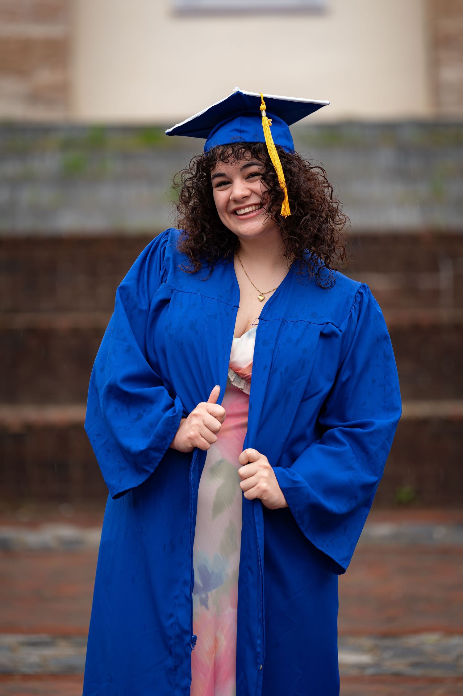 Graduate in blue cap and gown smiles while posing on brick steps.