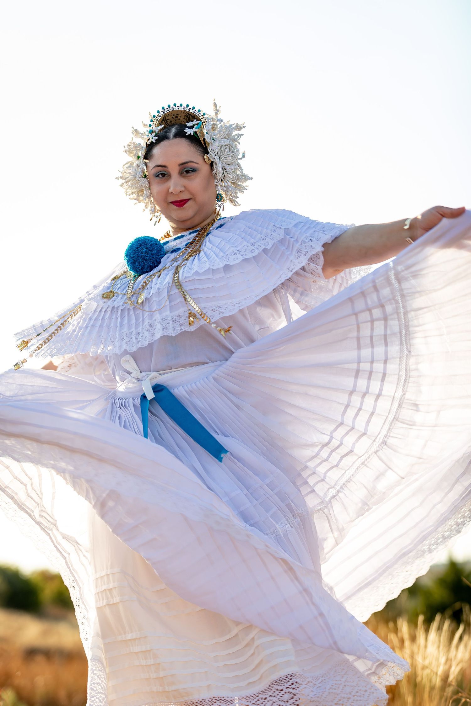 Dancer in white dress with blue sash twirls gracefully against bright natural backdrop.
