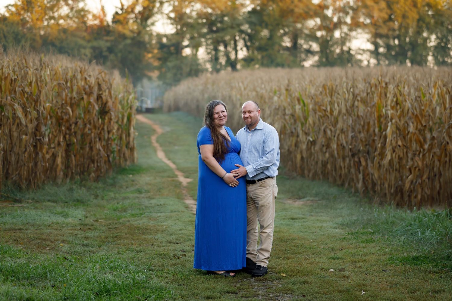 A couple embraces in a cornfield during golden hour with autumn trees in the background.
