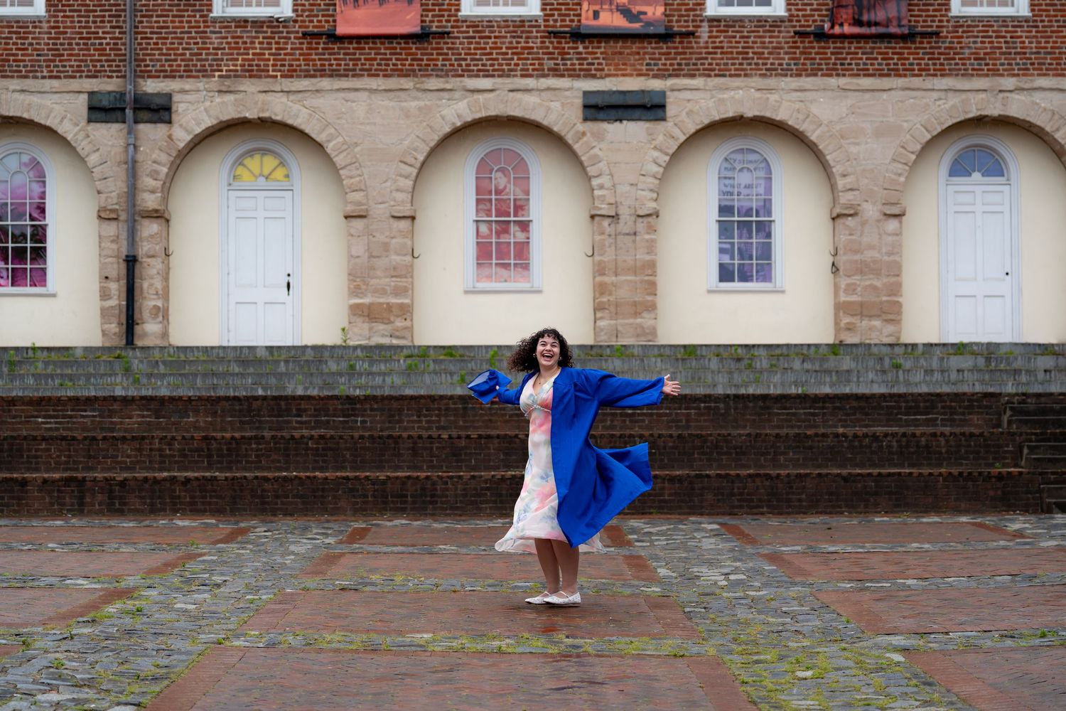 Graduate in blue gown twirls on brick steps in front of historic building with colorful doors.