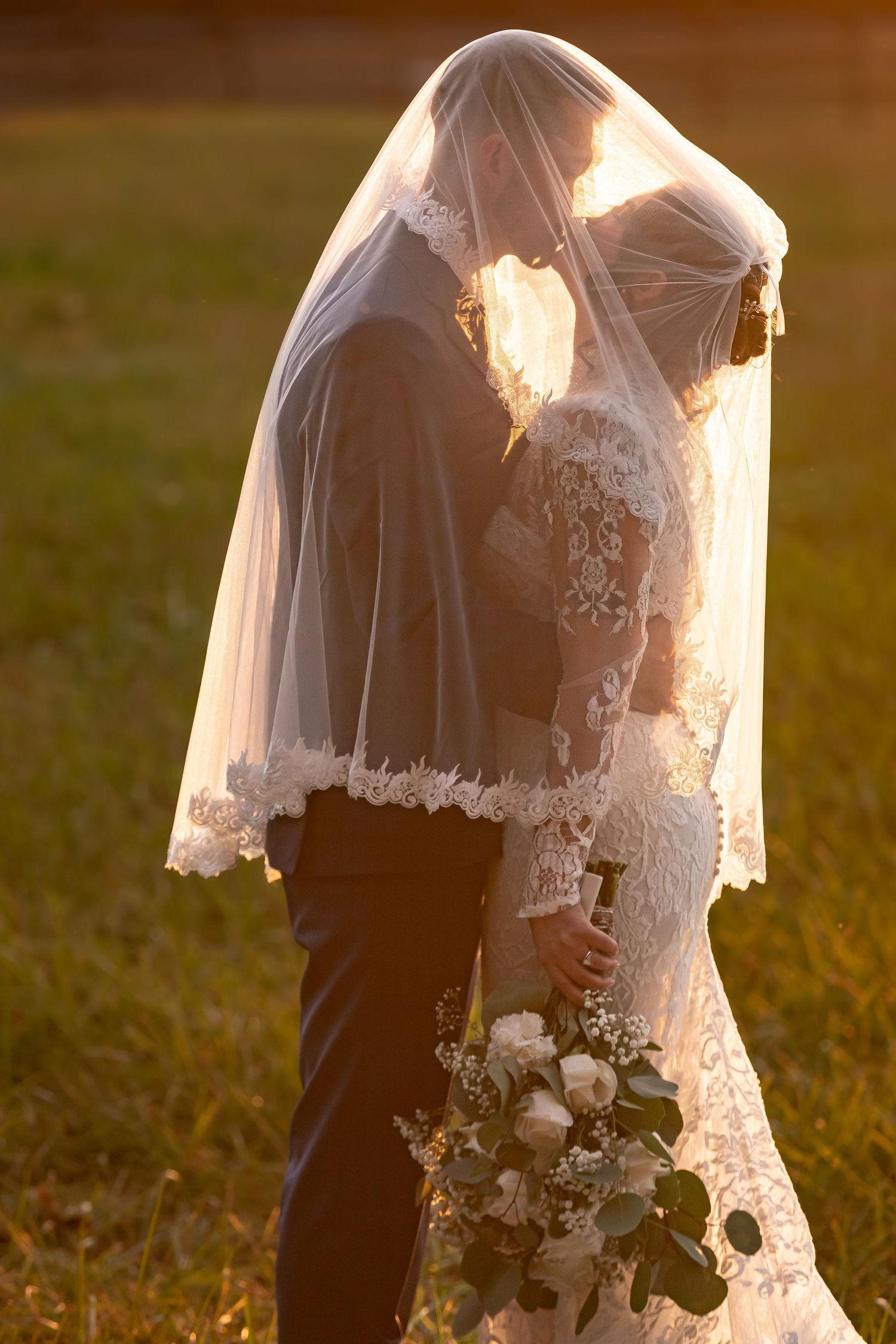 Romantic sunset silhouette of bride and groom with lace veil and white flowers.
