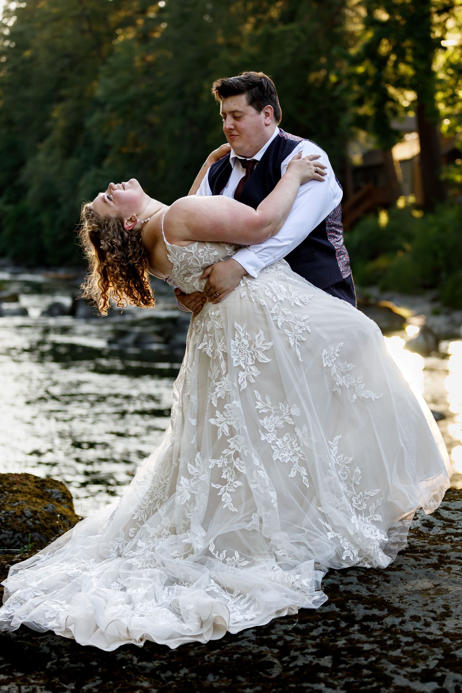 Romantic wedding couple performs dramatic dips during sunset photo shoot along a riverbank.