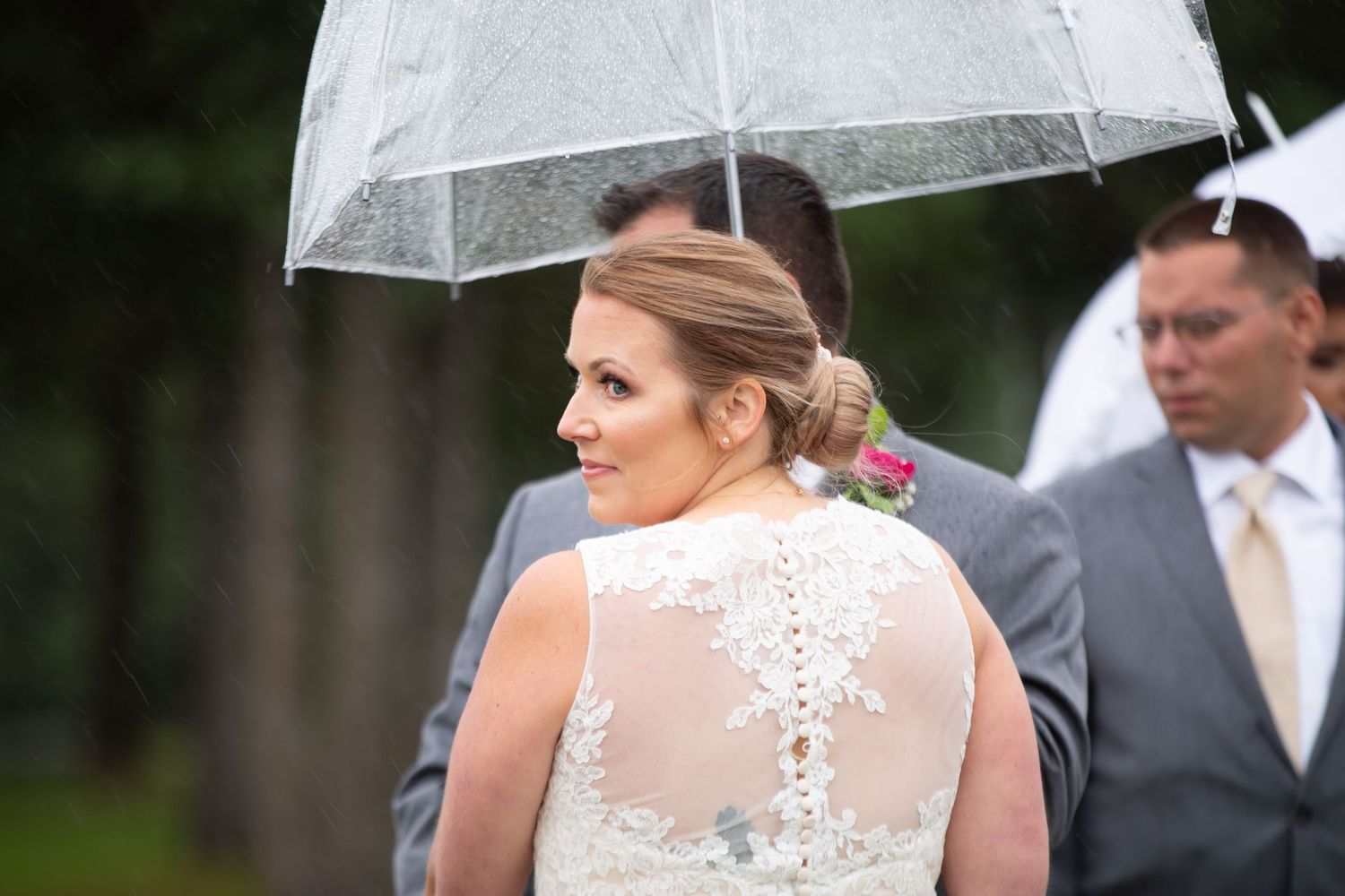 Person in elegant white wedding dress holding clear umbrella outdoors.