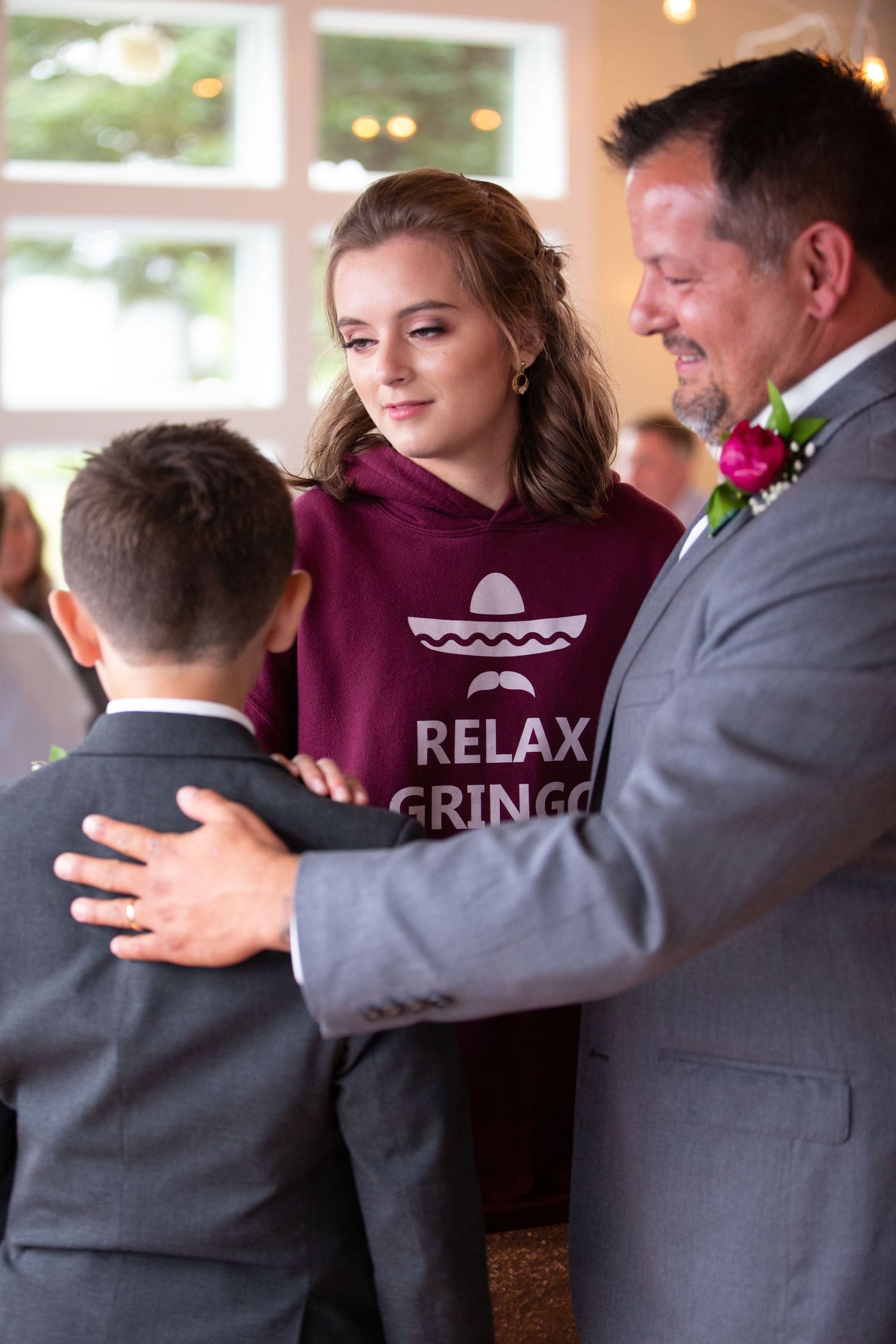 Wedding party members wearing burgundy and grey outfits share emotional hugs during reception celebration.
