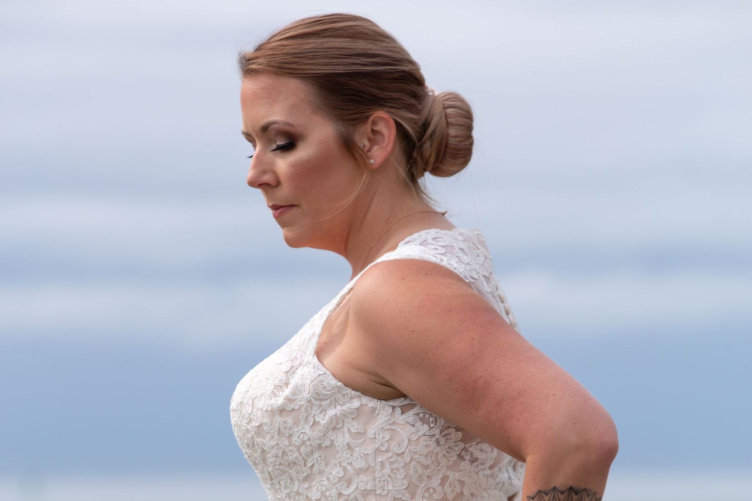 A bride in a white strapless wedding dress poses outdoors with a flowing veil against a cloudy sky.