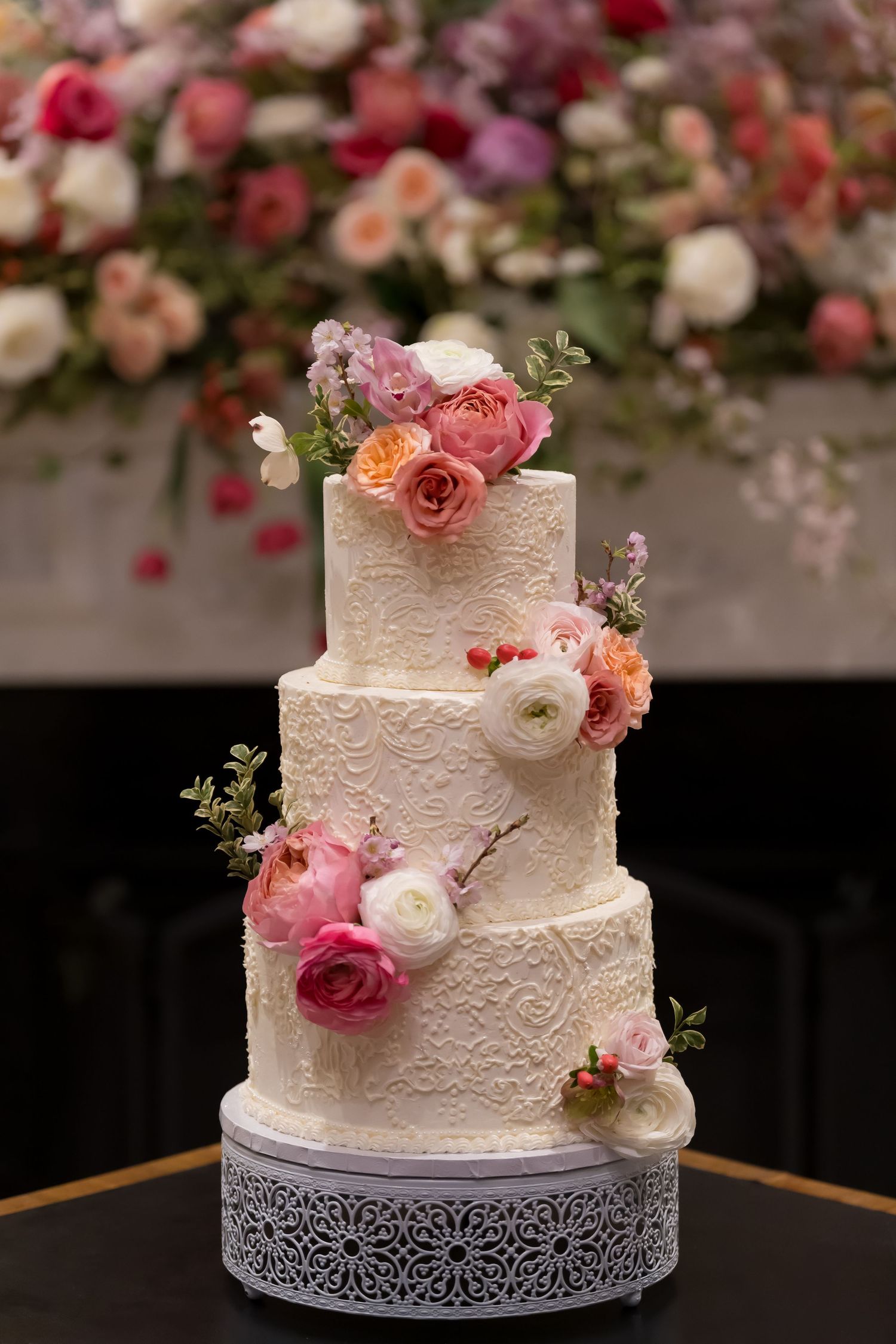 Elegant three-tier white wedding cake decorated with delicate lace patterns and fresh pink and coral roses on a silver stand.