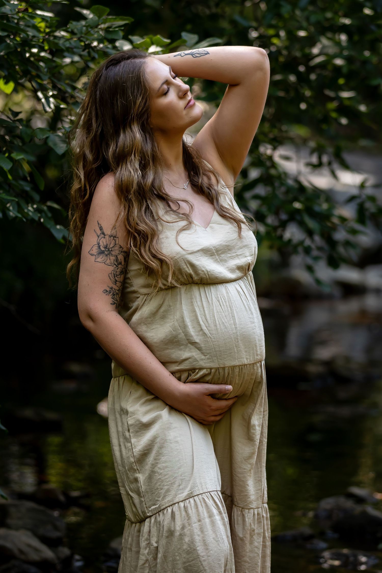 Artistic maternity portrait by water featuring flowing beige dress and natural scenery.