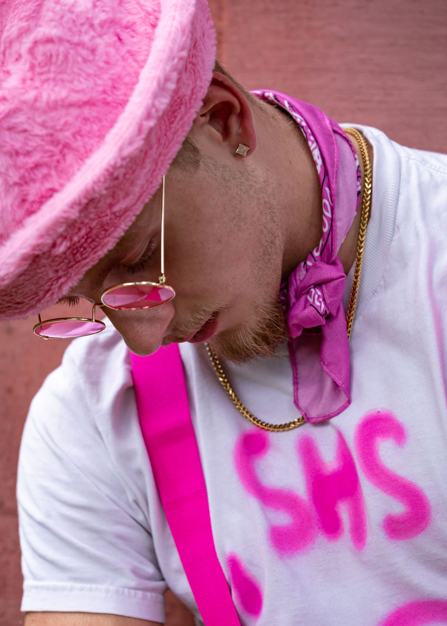 Close-up profile showing pink fuzzy hat, bandana and earring against brick background.