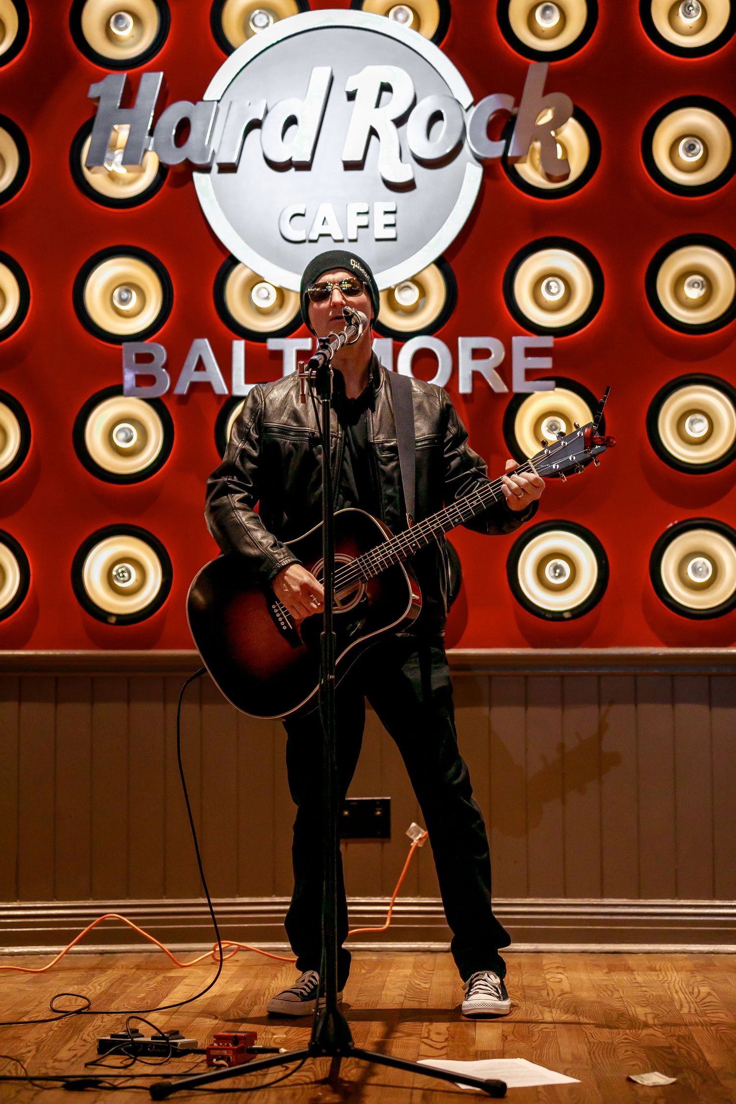 Musician performs with an acoustic guitar on stage at Hard Rock Cafe Baltimore with decorative lighting in background.