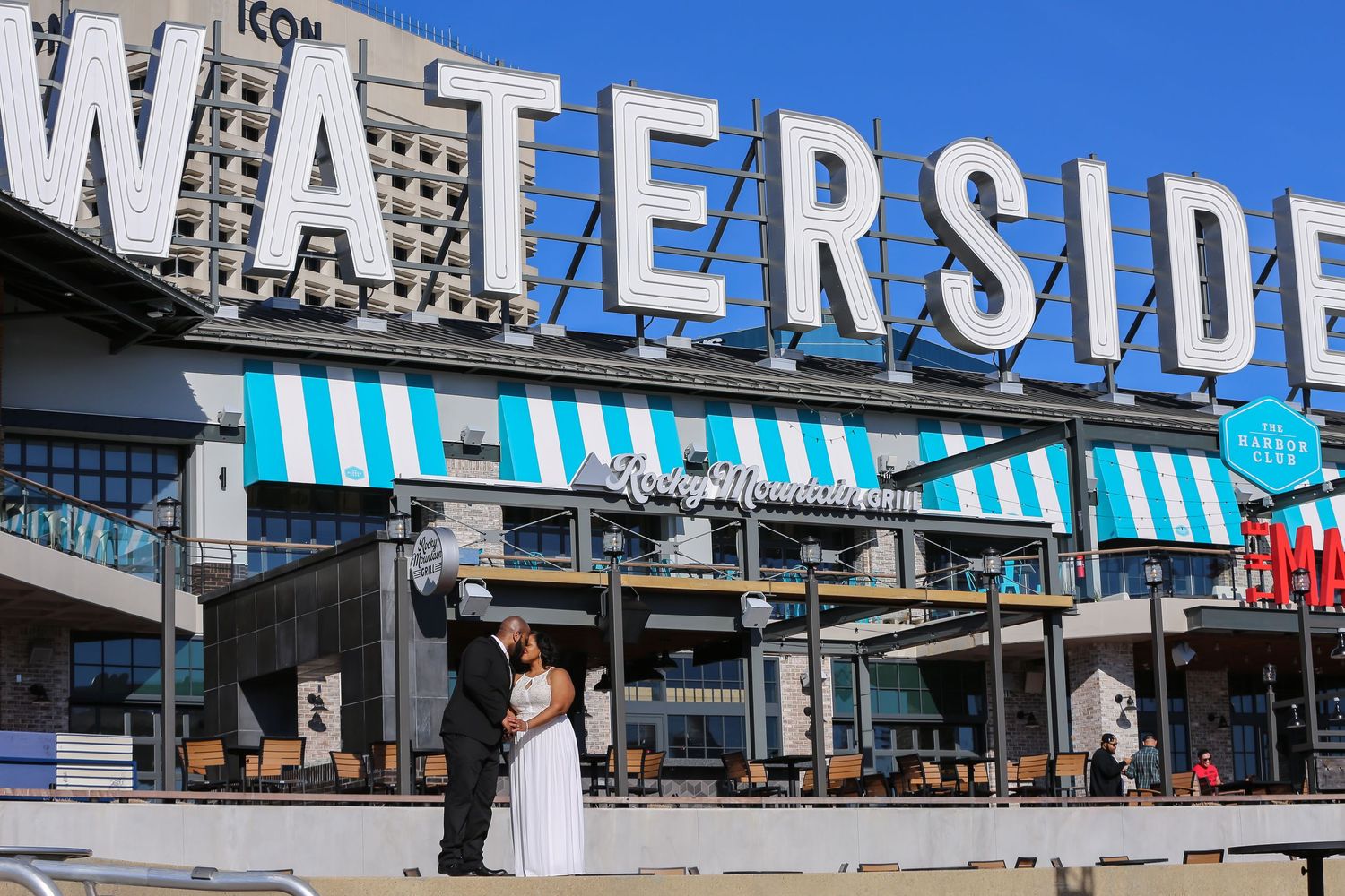 Waterside shopping and dining complex exterior with blue signage against bright sky and outdoor seating area.