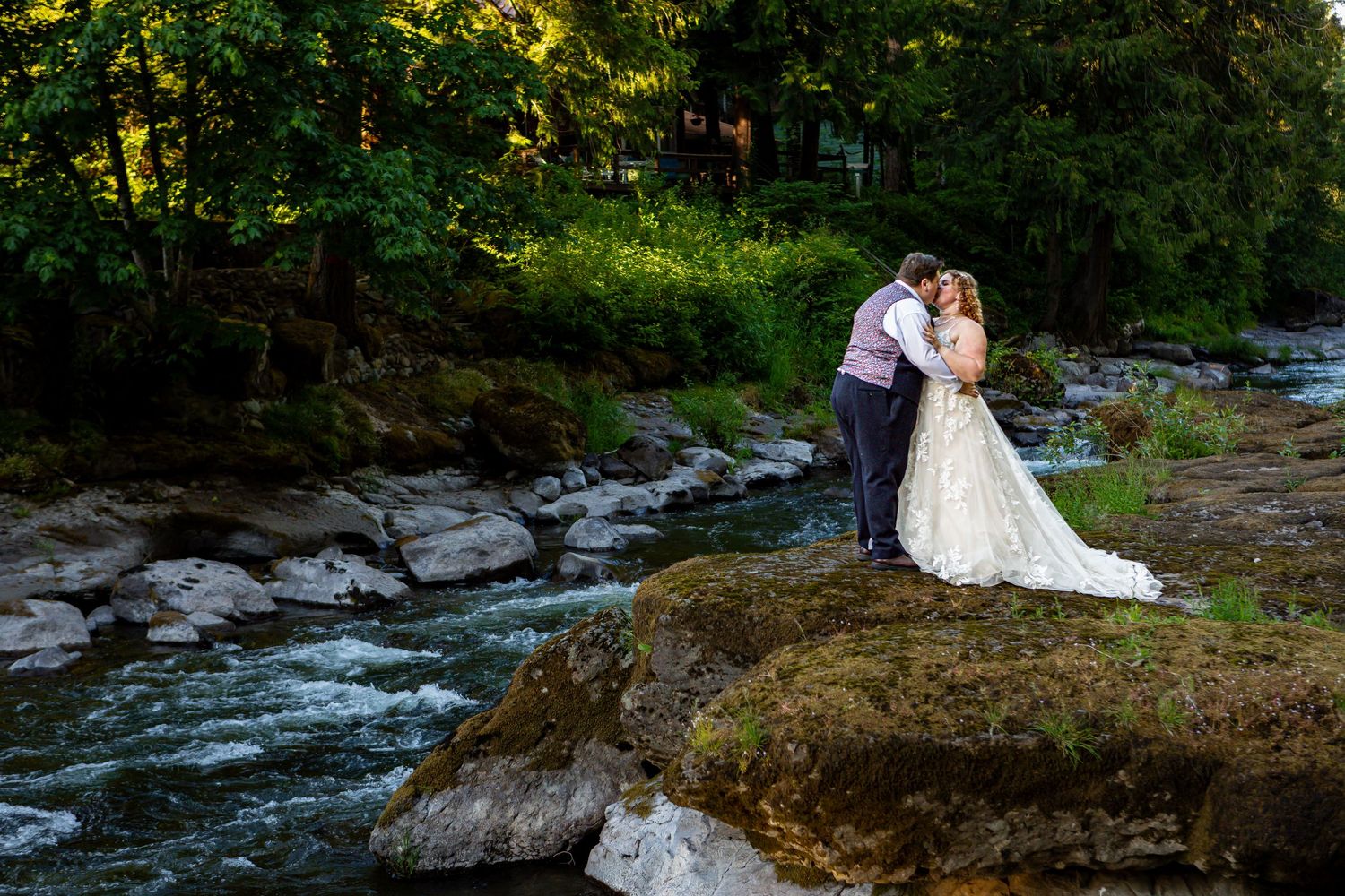 Newlyweds pose for romantic photos beside a rushing river in a forest setting.