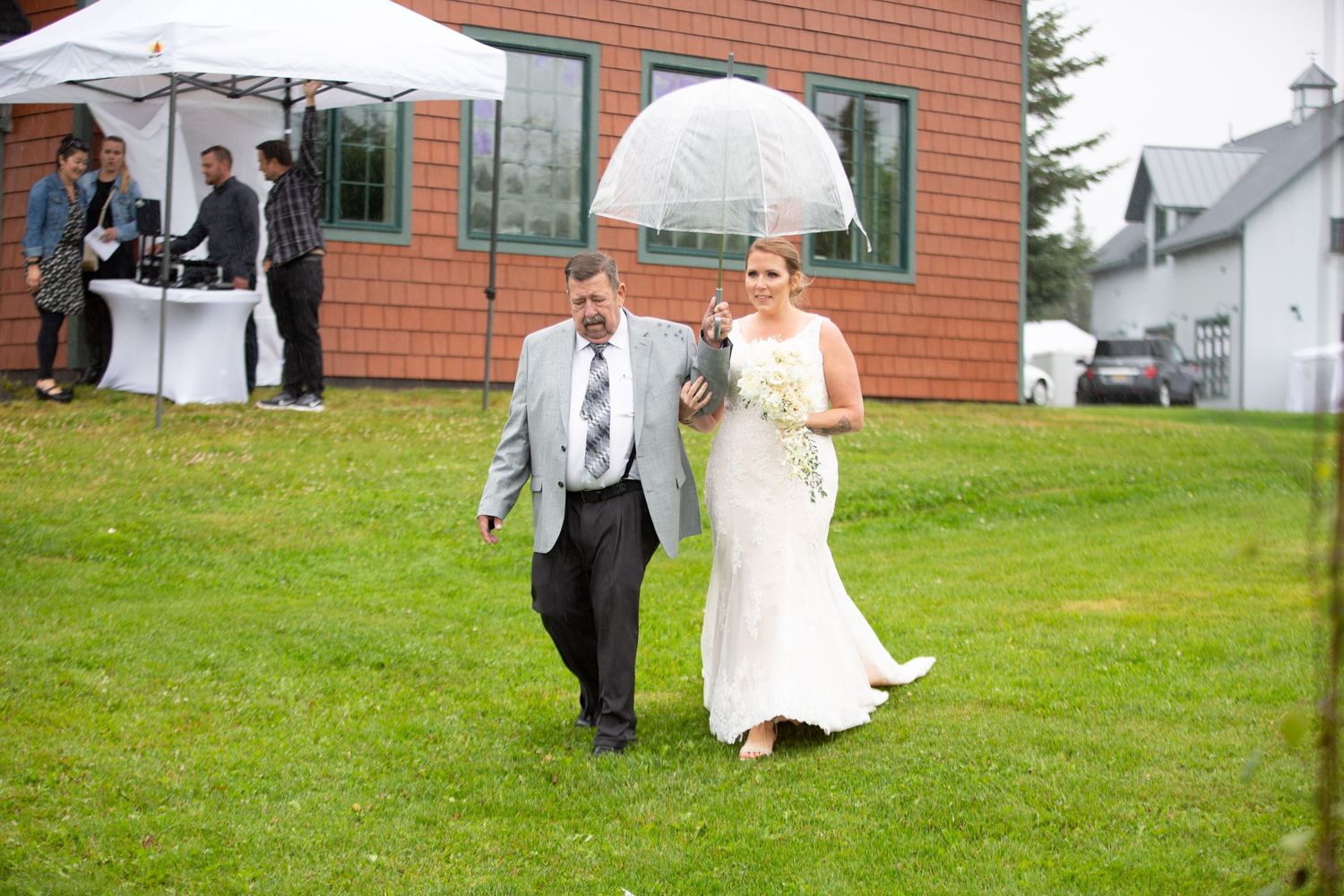Wedding party walking outdoors with white tents and brick building in background.