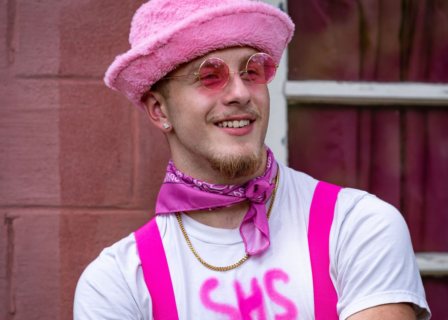 Person wearing pink fuzzy hat and suspenders with rose-tinted glasses against brick wall.