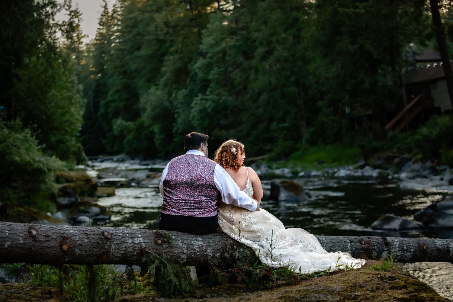 A couple sits peacefully on a log overlooking a serene forest stream.