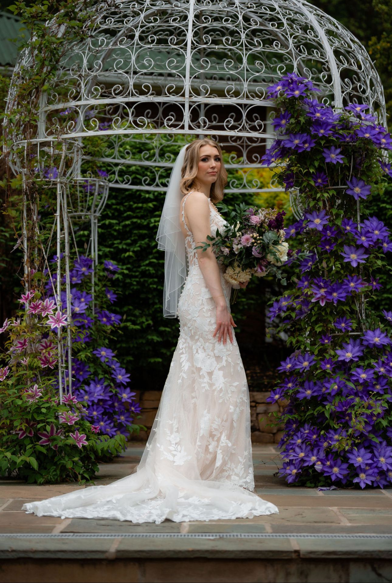 Beautiful white lace wedding dress photographed in a garden gazebo with vibrant purple clematis flowers blooming.