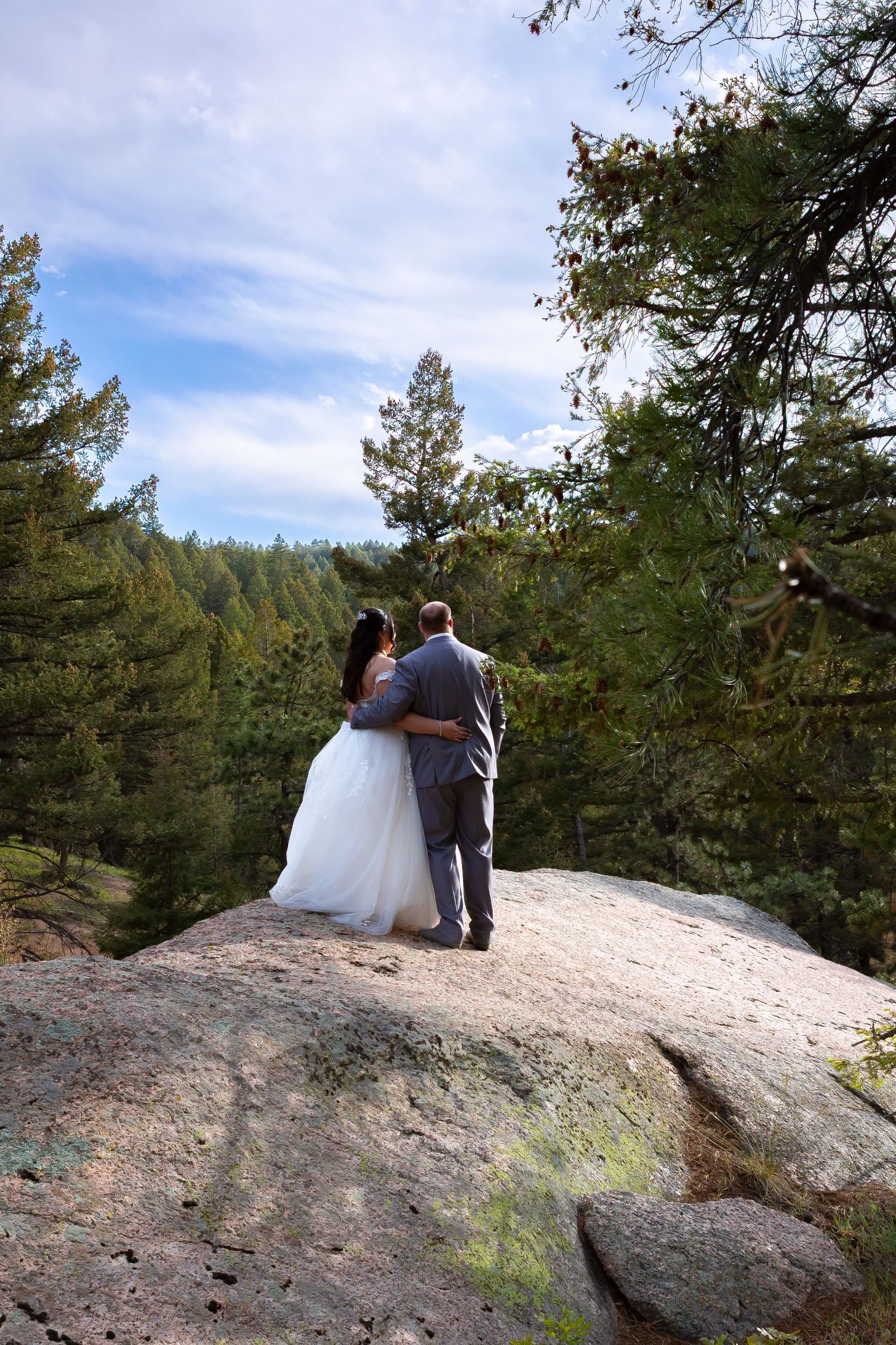 A couple stands together on a scenic rocky overlook surrounded by pine trees.