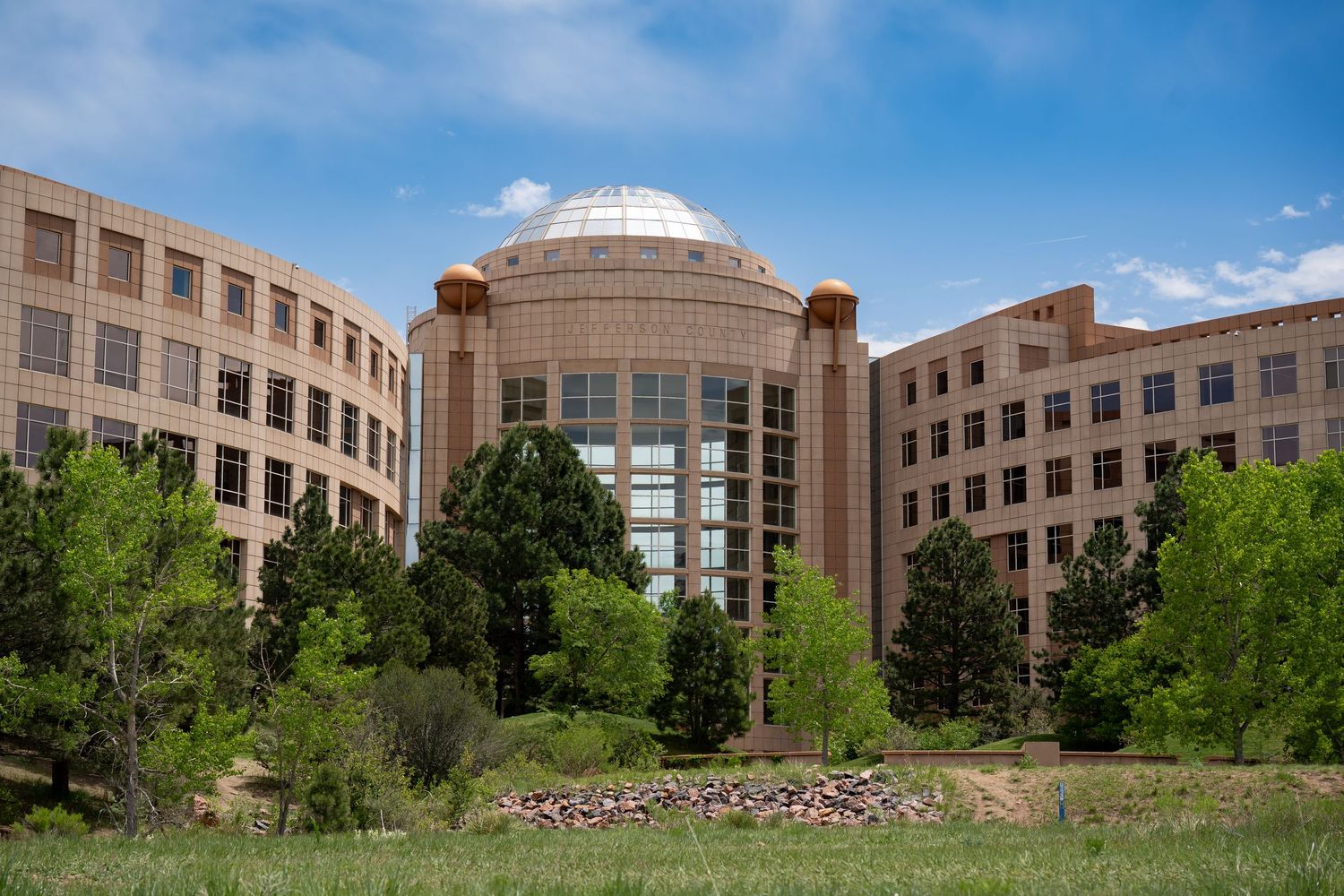 Modern government building with domed center and symmetrical wings surrounded by trees.
