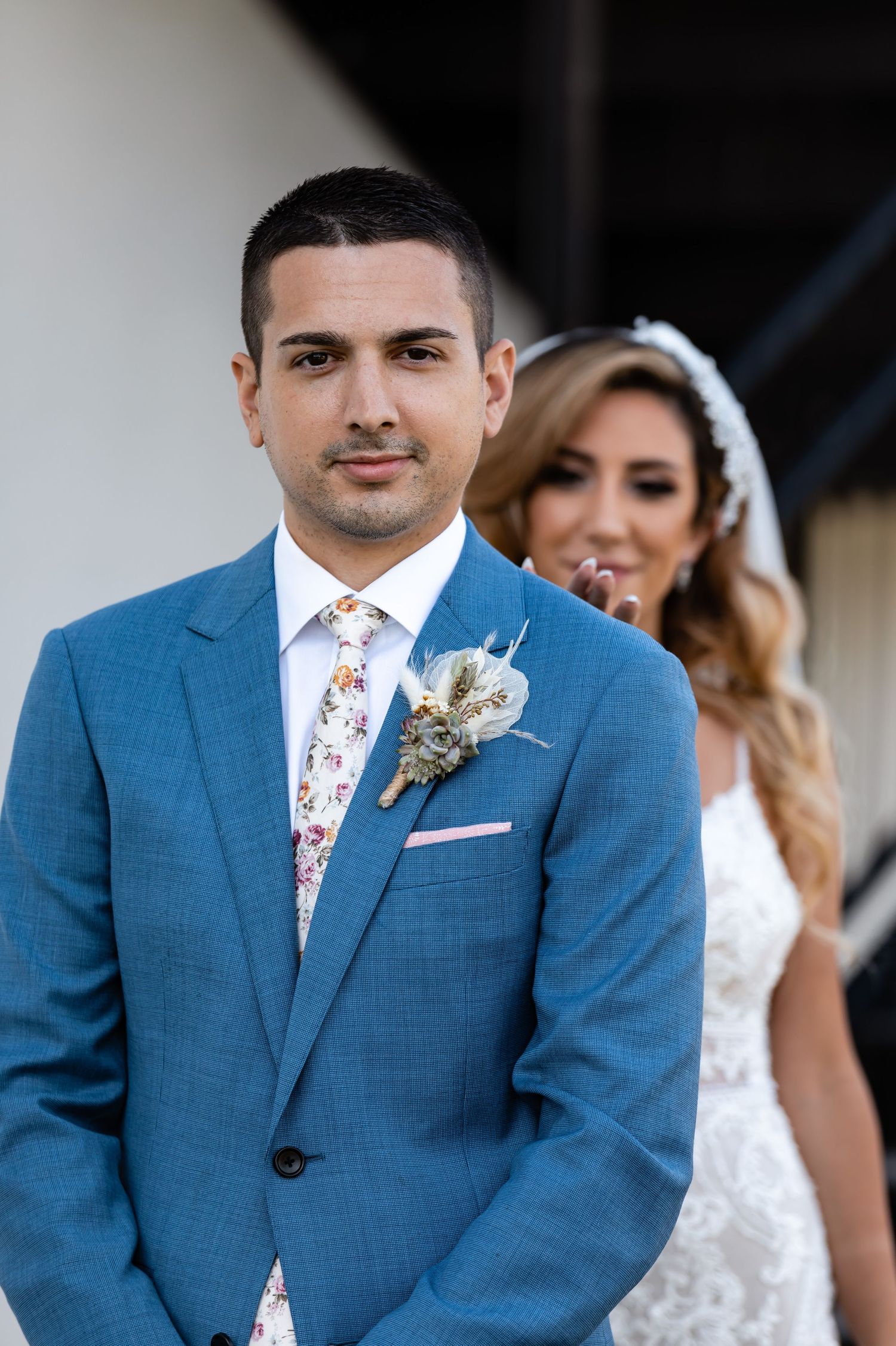 Groom in blue suit with floral tie and boutonniere against neutral background.