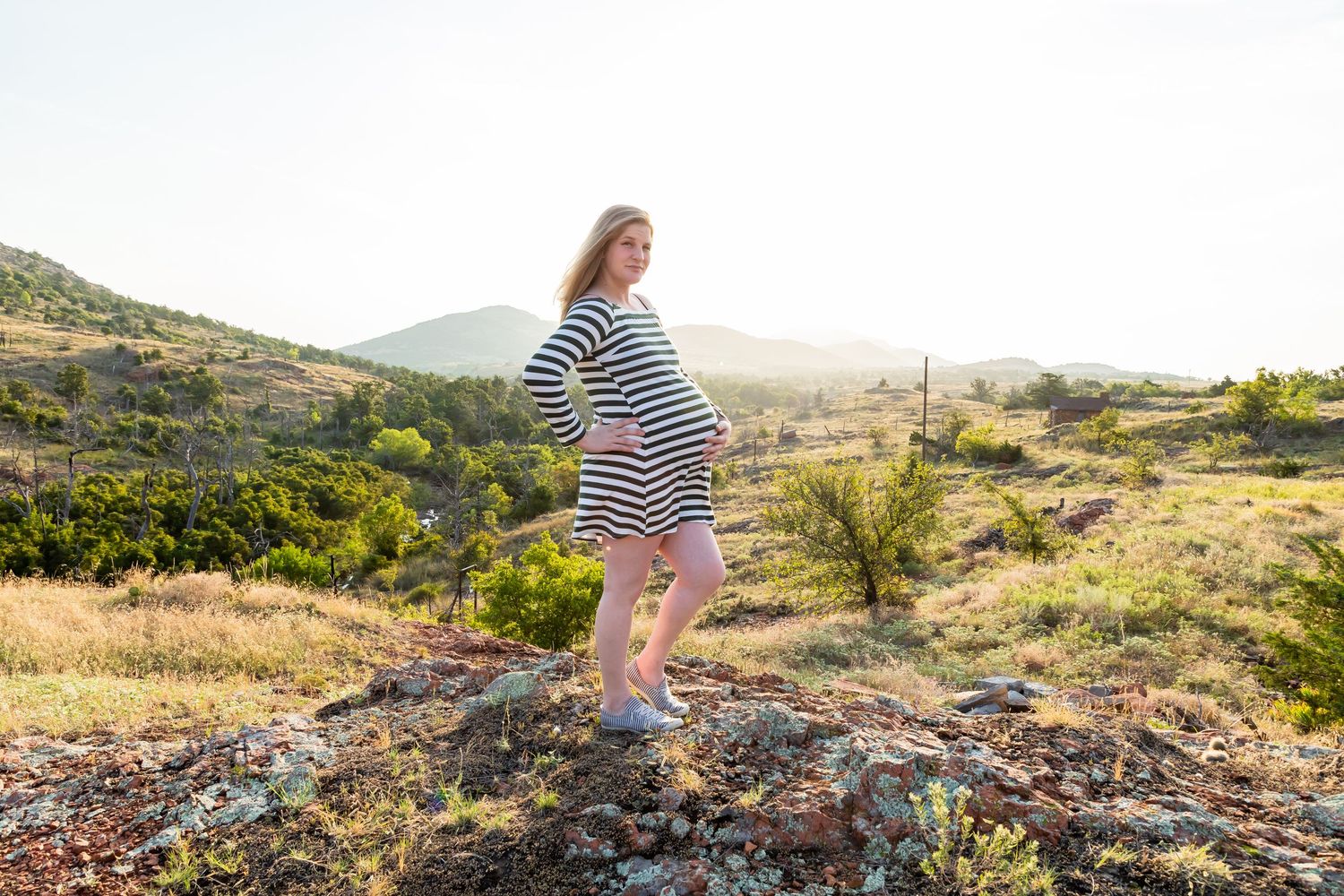 Maternity photo showing a woman in striped dress standing on rocky terrain at sunset.