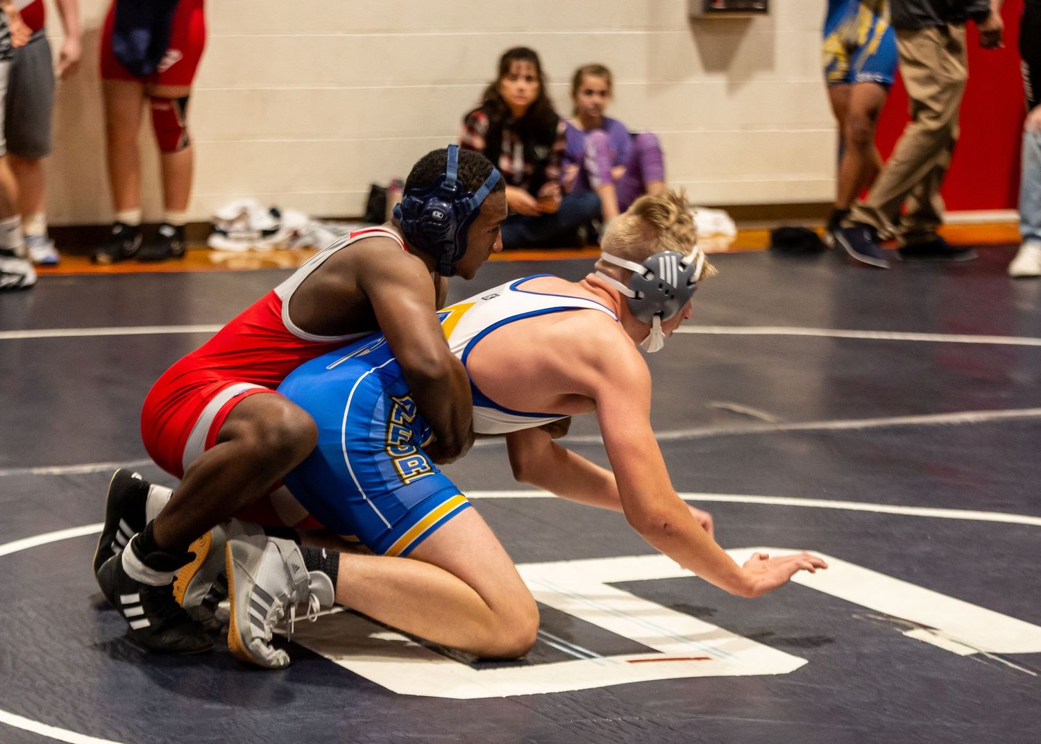 Close-up of wrestlers grappling on black mat during indoor competition.