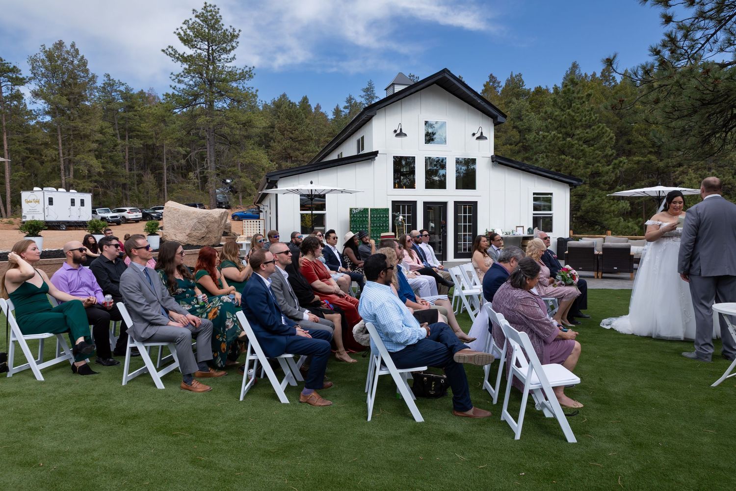 A panoramic view of wedding guests seated on white chairs with a white building and pine trees in the background.