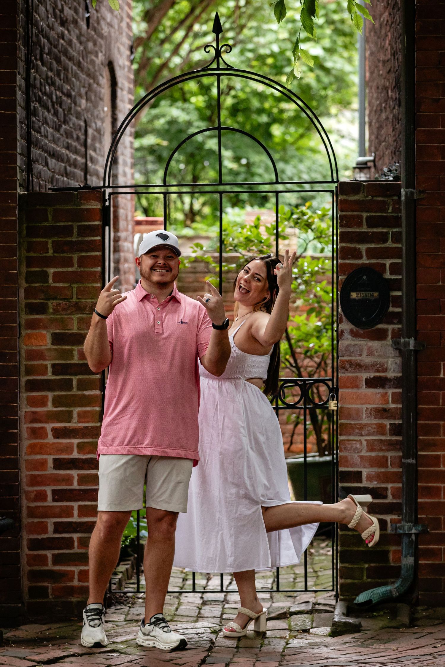 Two people pose playfully by an ornate iron archway against a backdrop of historic brick architecture.