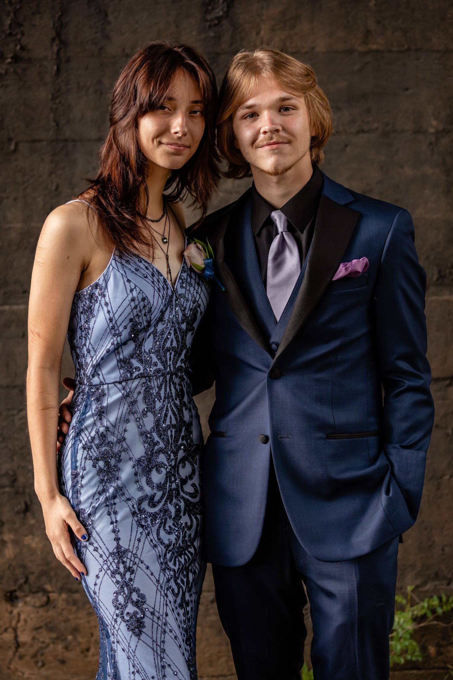 A couple in formal wear poses together against a textured wall.