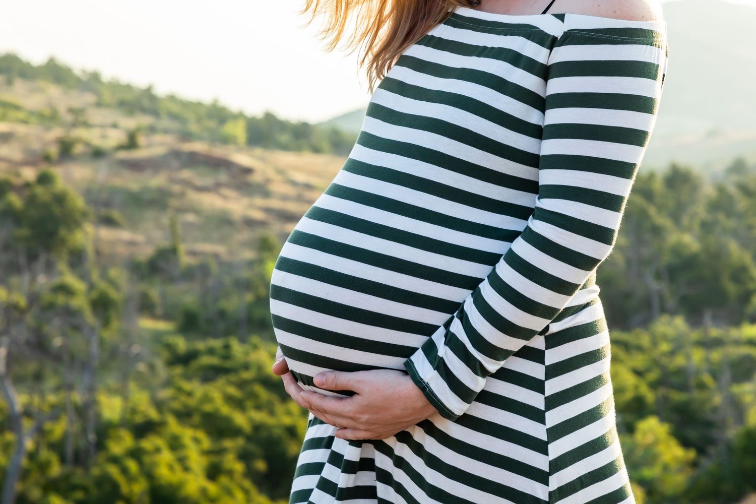 Close-up of a pregnant belly in a striped shirt against a natural outdoor backdrop.