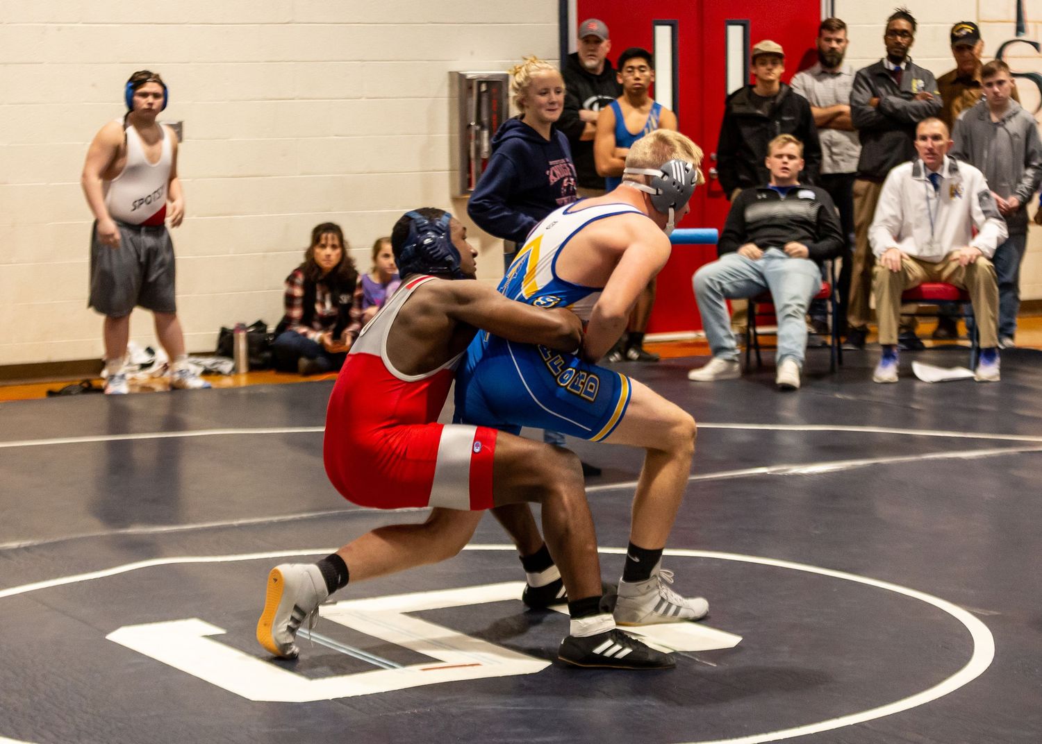 Wrestling competition in progress on indoor mat with spectators watching from the sides.