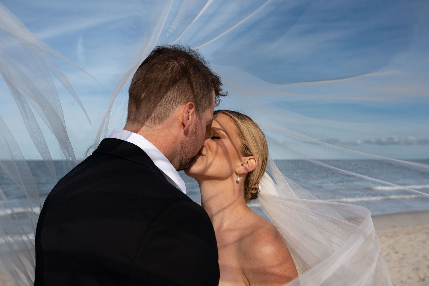 Romantic beach wedding photo with flowing veil against blue sky.