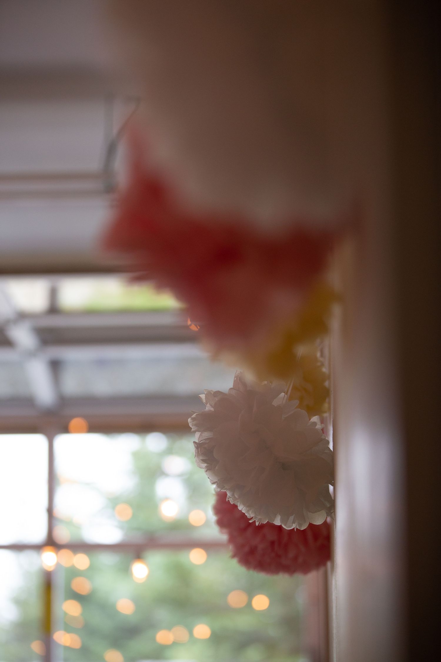 Decorative white and red tissue paper pom poms hanging near window with bokeh lights.