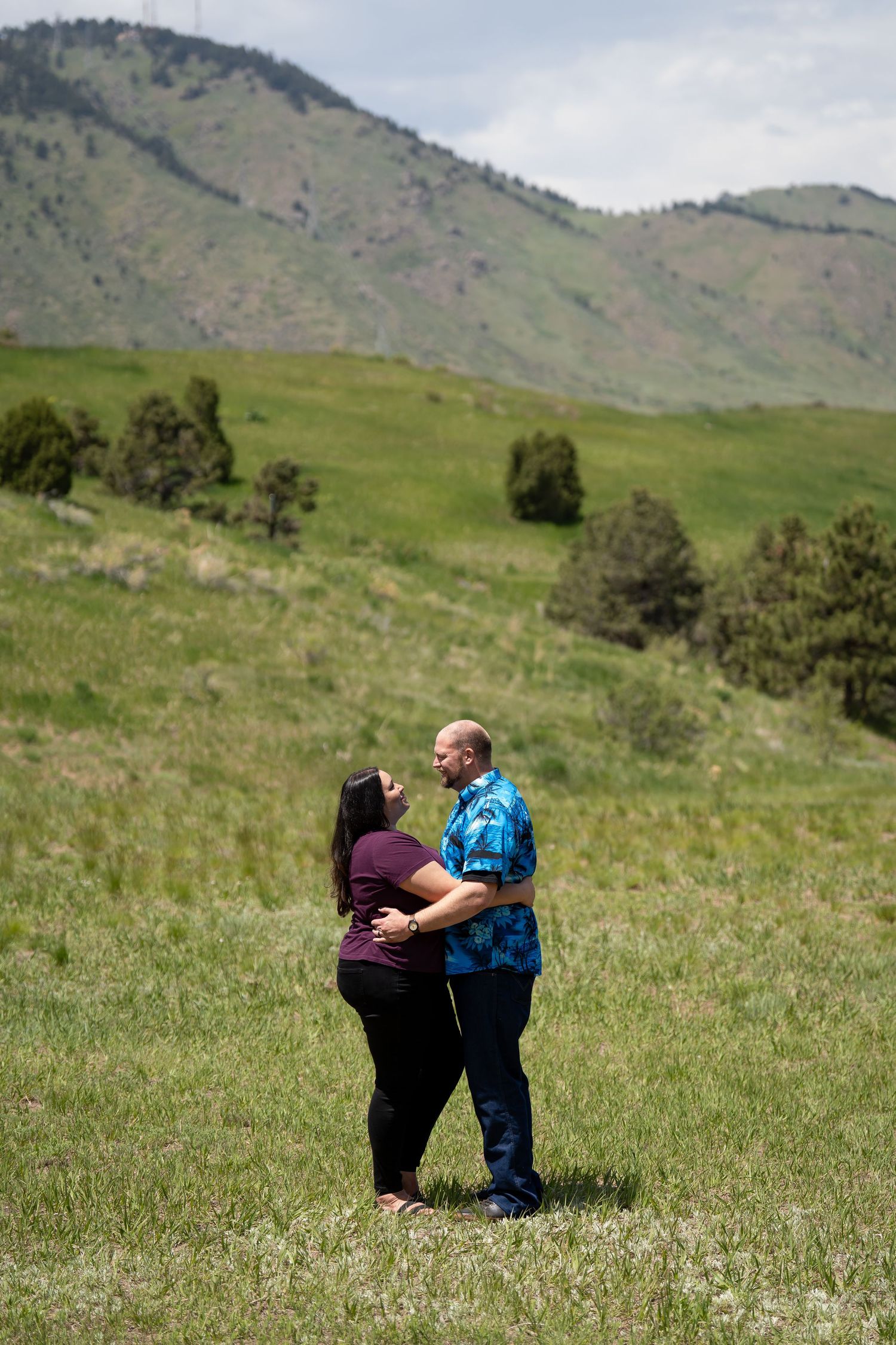 A couple embraces in a scenic mountain meadow with green rolling hills in the background.